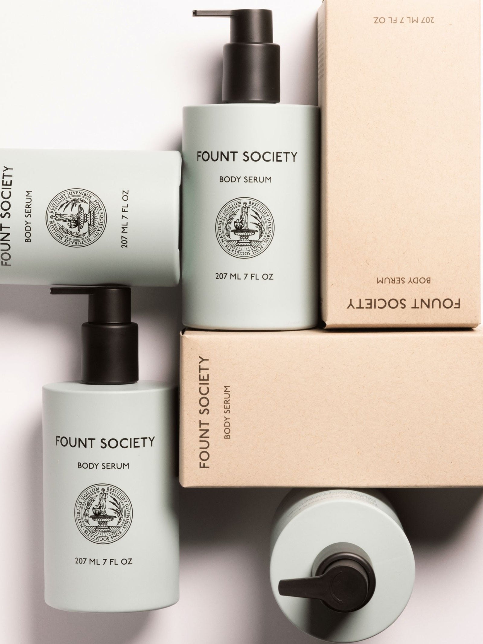 Arrangement of Cozy Earth Body Serum bottles, each 207 ml/7 fl. oz., with minimalist labels featuring a circular emblem. Some bottles lie horizontally while others stand upright. Two cardboard boxes are also included. The scene is bright and clean.