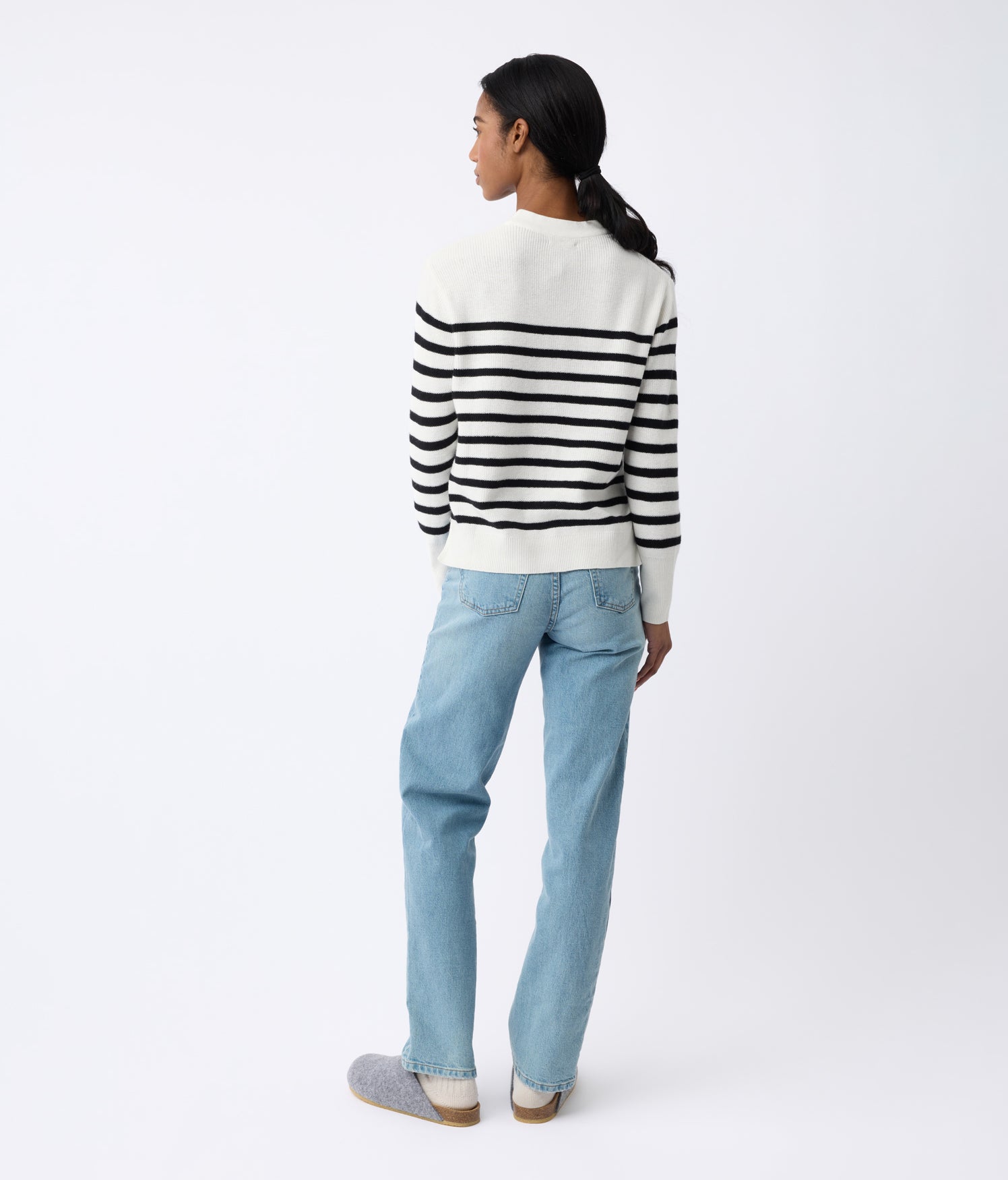 A person is standing, facing away, wearing a Cozy Earth Women's Rowan Sweater with black stripes, light blue jeans, and gray slip-on shoes. The background is plain and the person has their hair in a low ponytail. |Color:Bone/Jet Black Stripe