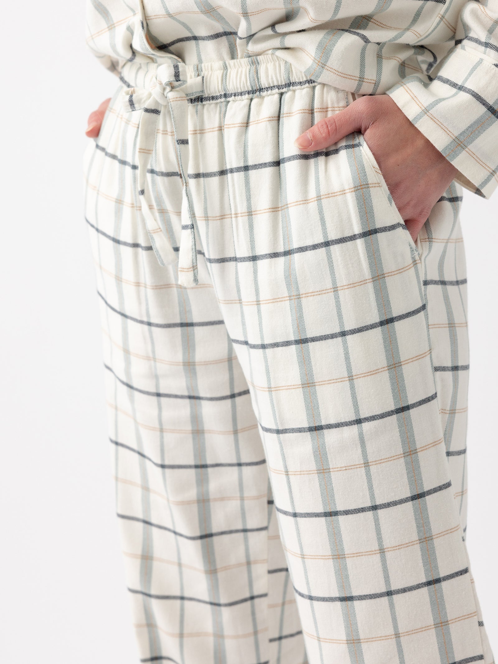 A person is pictured from the waist down, wearing the Women's Farmhouse Long Sleeve Pajama Set from Cozy Earth. The pajama set has a white base with a blue and beige grid pattern. Their hands rest in the pockets of the pants, which feature an elastic waistband and a drawstring tie. The background is plain white. 