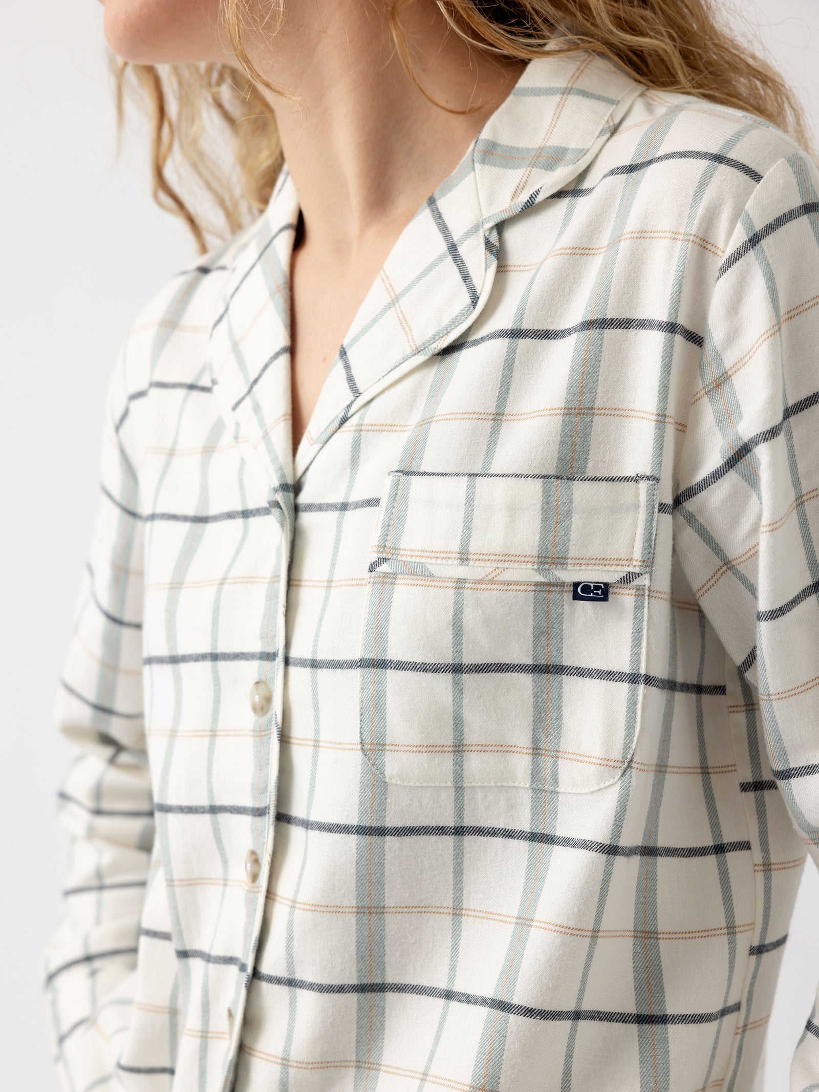 A person with wavy blond hair is partially visible, wearing the Women's Farmhouse Long Sleeve Pajama Top by Cozy Earth. The top features a button-up design and a light-colored plaid pattern, along with a small chest pocket adorned with a logo tag. The background is plain and white. 