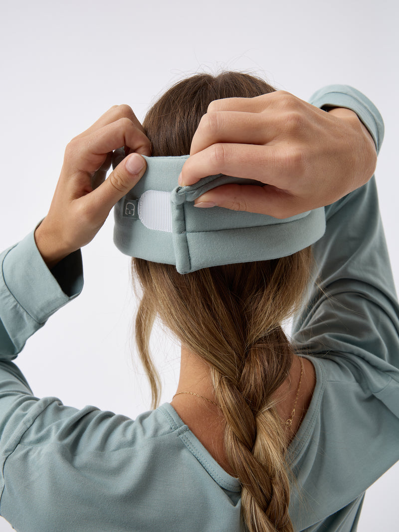 A woman wears the Cozy Earth Adjustable Bamboo Sleep Mask in Brook. She is facing away and showing where the mask fastens at the back.