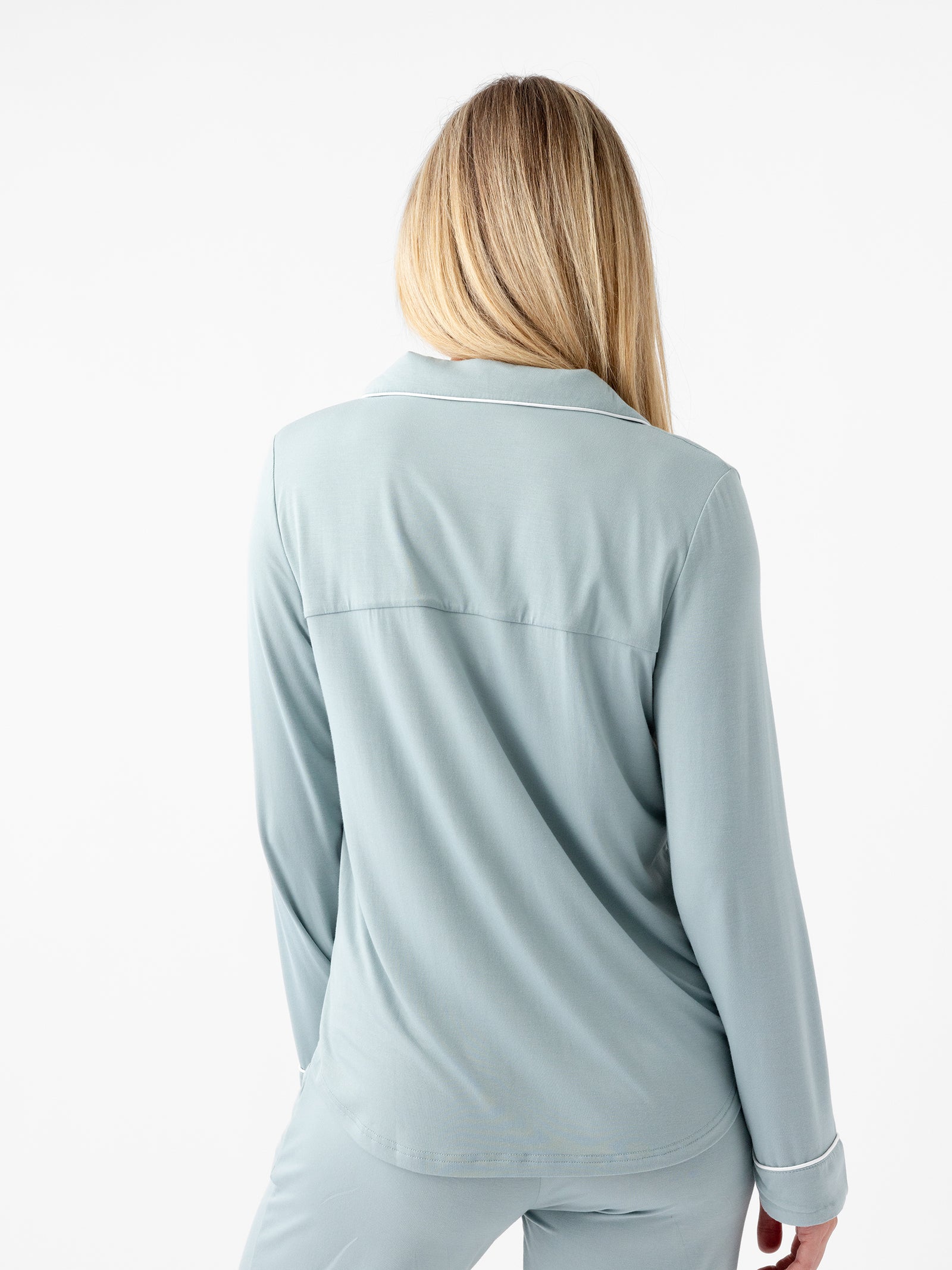 A person with long blonde hair is shown from the back, wearing the Women's Long Sleeve Bamboo Pajama Top in Stretch-Knit by HIDE. The light blue pajama set with a collared top looks soft and comfortable against a plain white background. 