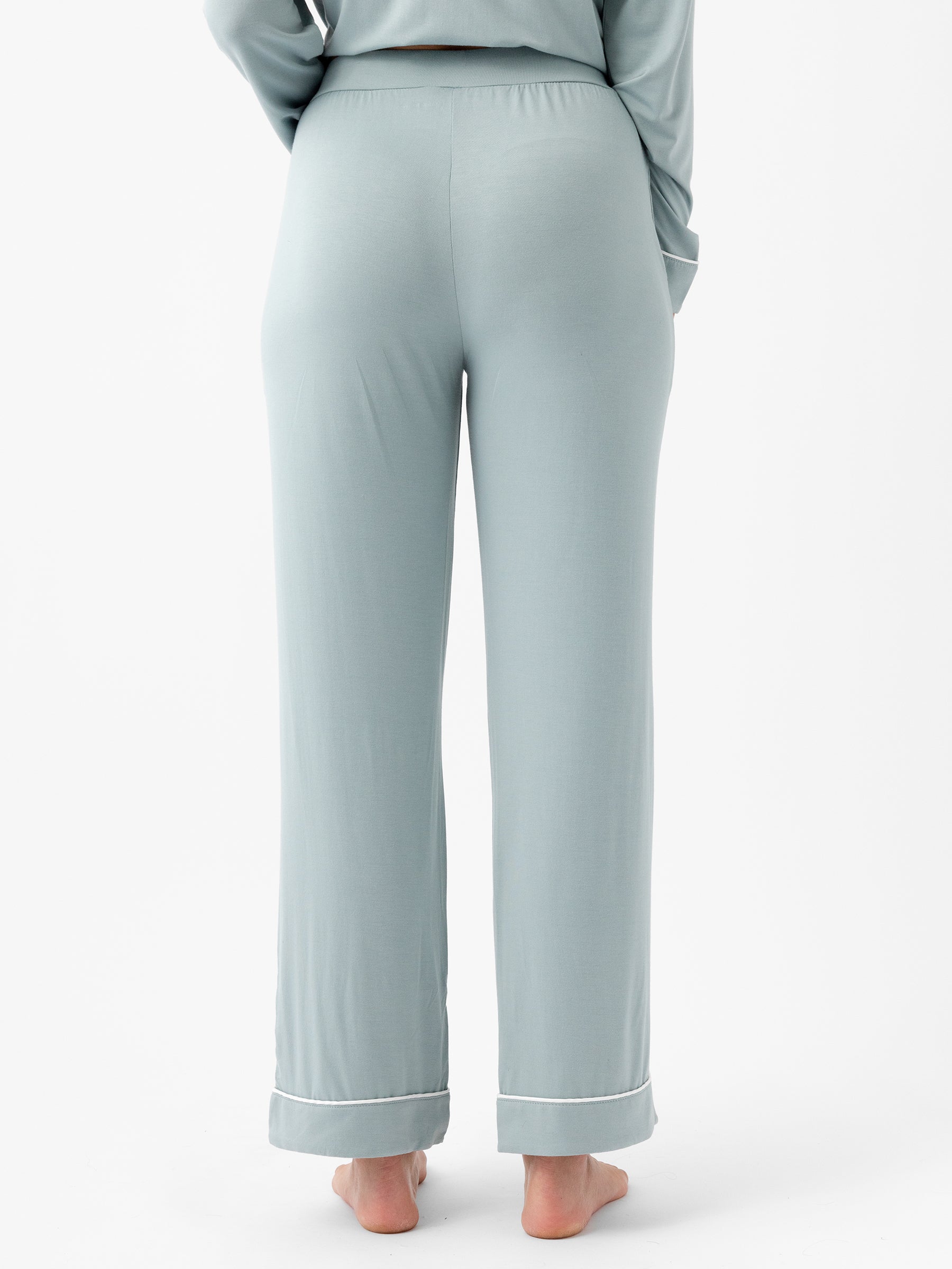 A person standing barefoot on a white background, wearing HIDE's Women's Stretch-Knit Bamboo Pajama Pant in light blue with white trim at the bottom. Only the lower body is visible, facing away from the camera. The pajama pants have a relaxed fit. |Color:Brook