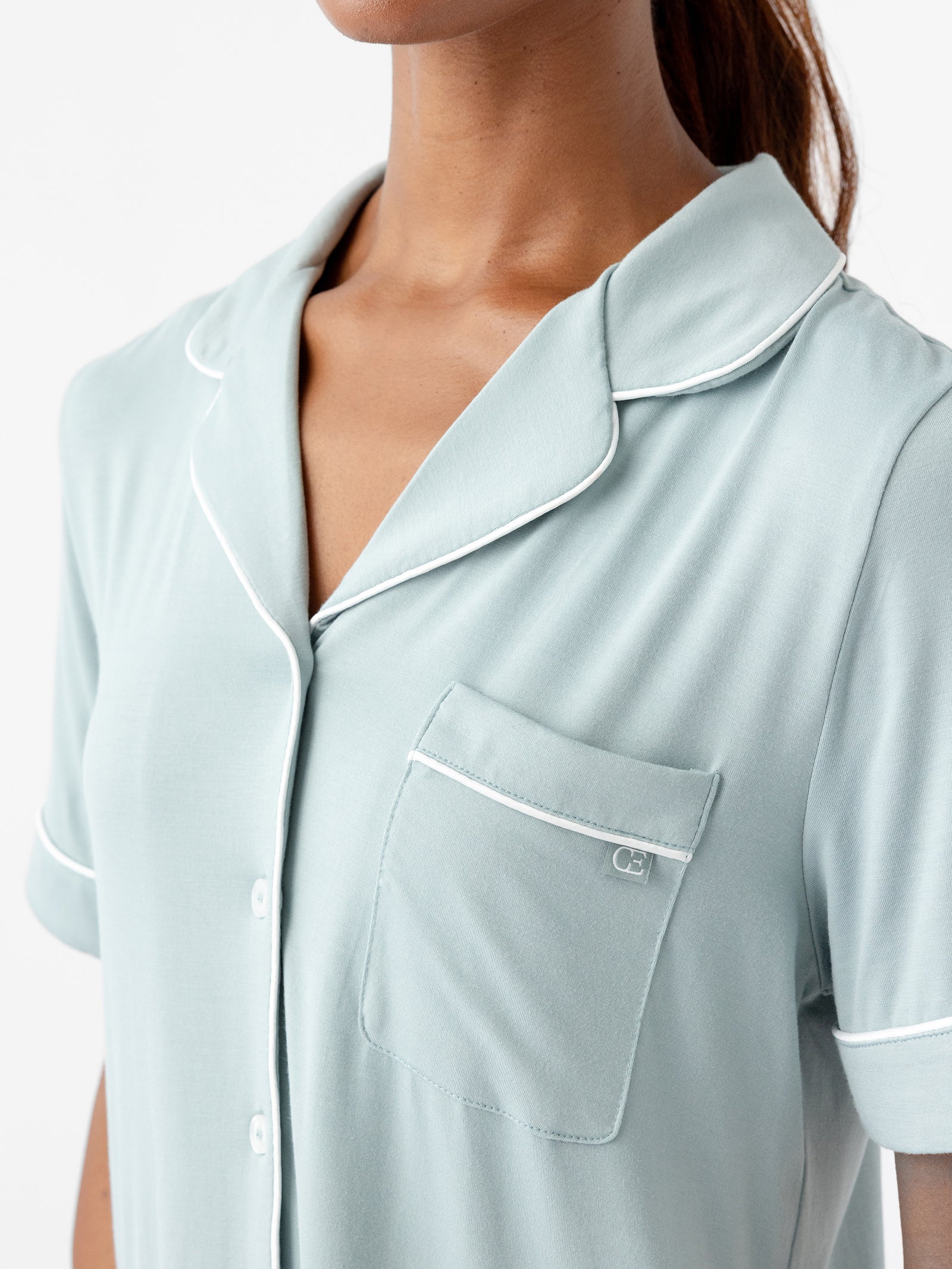 A close-up of a person wearing the Women's Short Sleeve Bamboo Pajama Top in Stretch-Knit by HIDE, featuring a light blue hue with white piping around the collar, pocket, cuffs, and button placket. The fabric appears soft and comfy, and a small logo is visible on the chest pocket. The person has their hair tied back. 