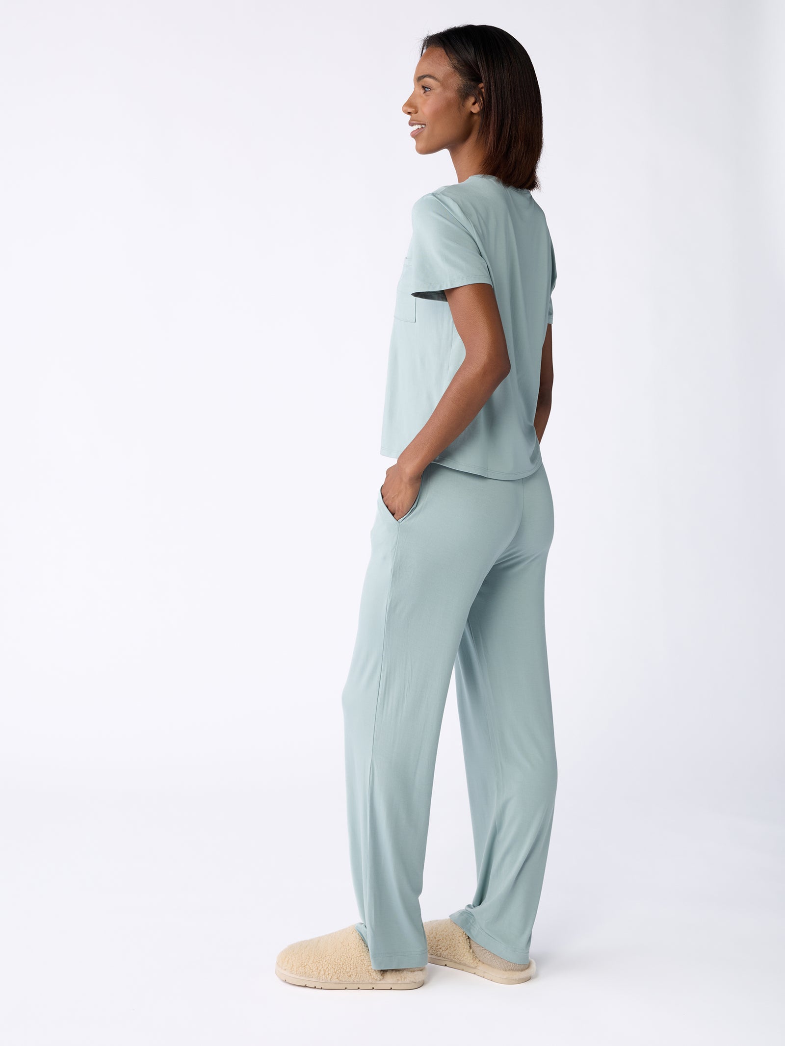 Wearing the Stretch Knit Bamboo Pant from Cozy Earth in light blue, paired with beige slippers, a person stands sideways against a plain white background. The ensemble looks comfortable and casual, perfect for lounging. 