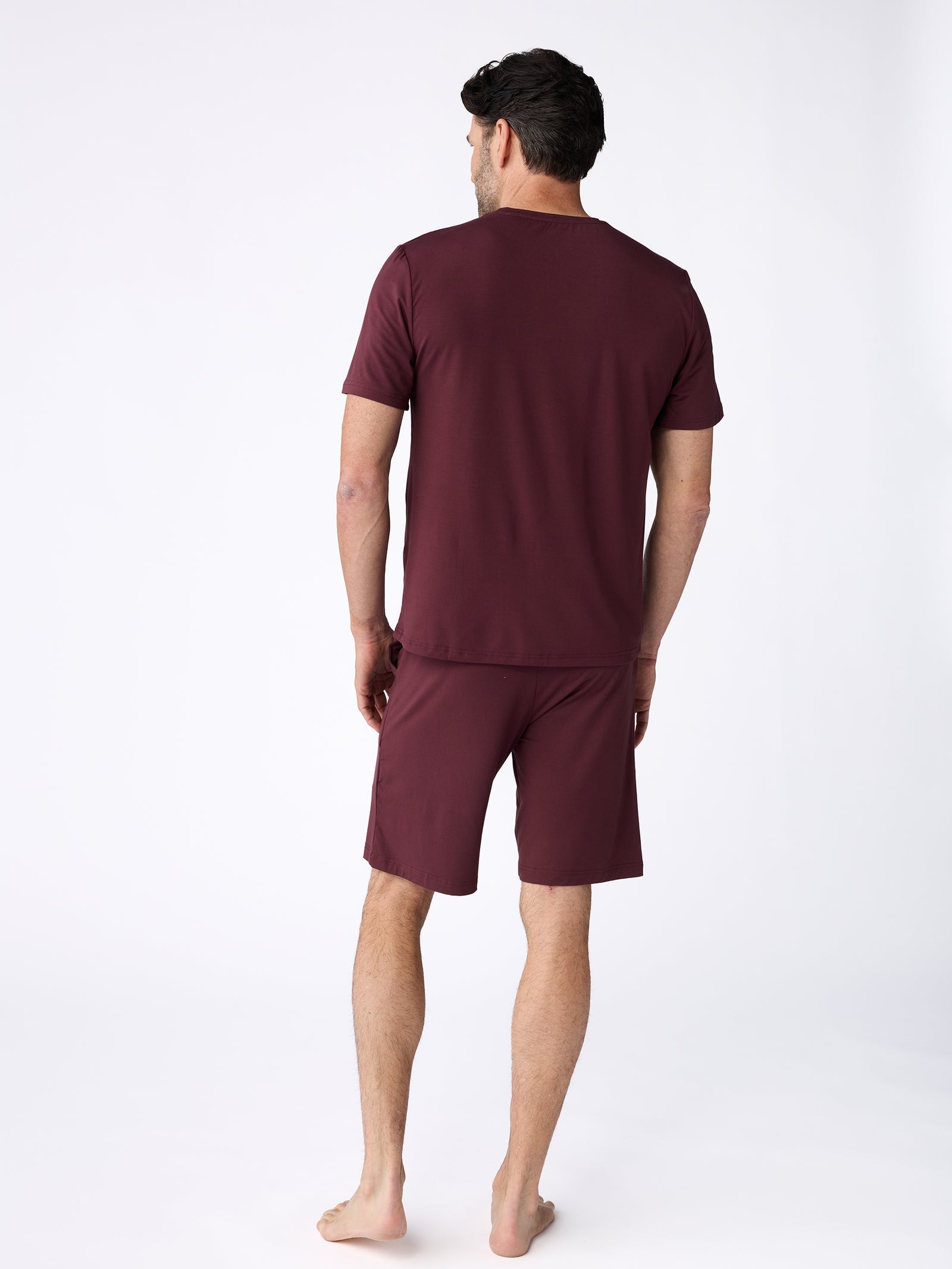 A person stands barefoot, facing away and wearing a matching maroon ensemble consisting of a Cozy Earth Men's Stretch-Knit Bamboo Pajama Short. They are on a plain white background. 