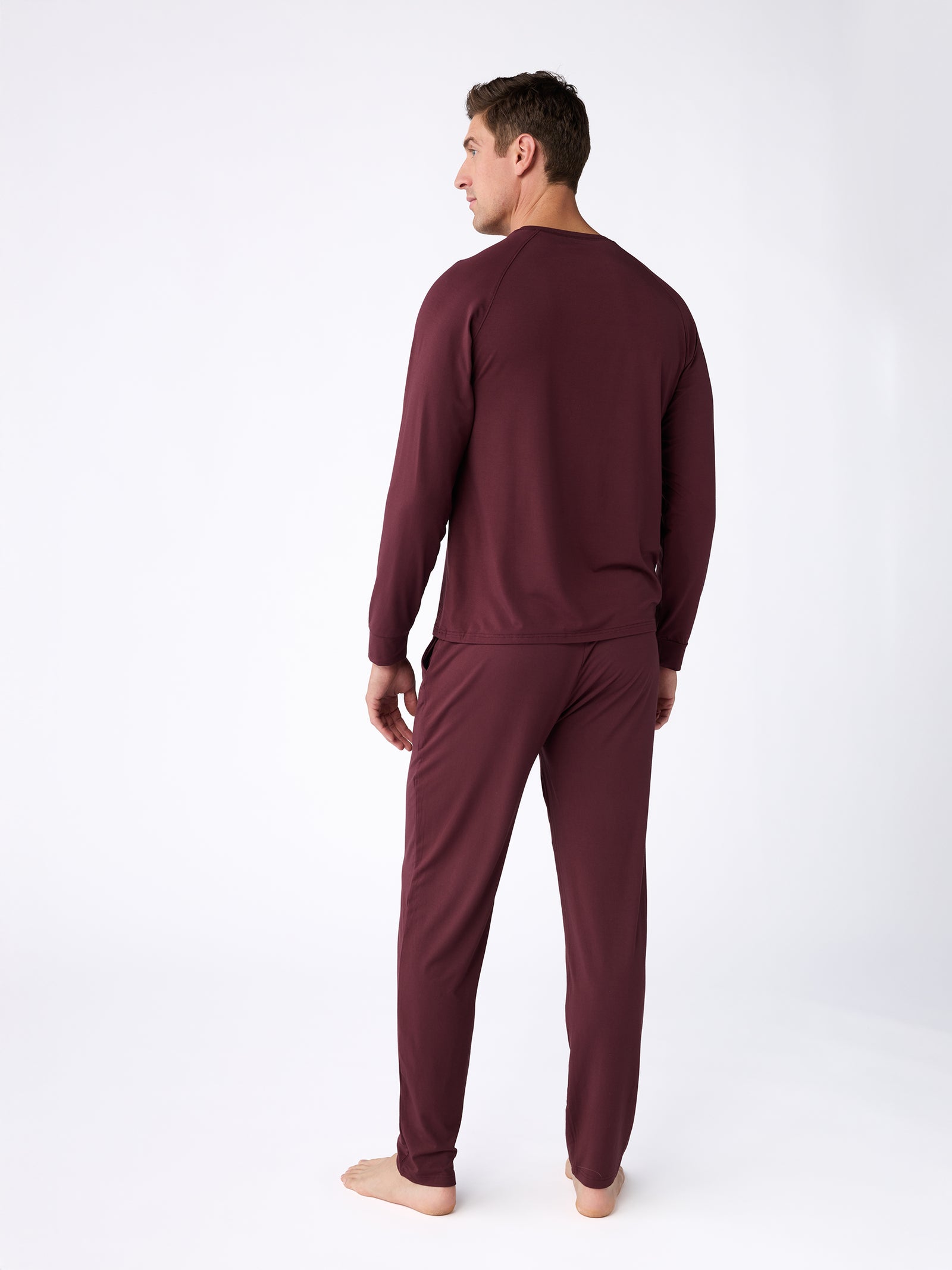 A man stands barefoot on a white background wearing Cozy Earth's maroon Men's Stretch-Knit Bamboo Pajama Pant set. He is facing away from the camera, highlighting the back of the outfit. 