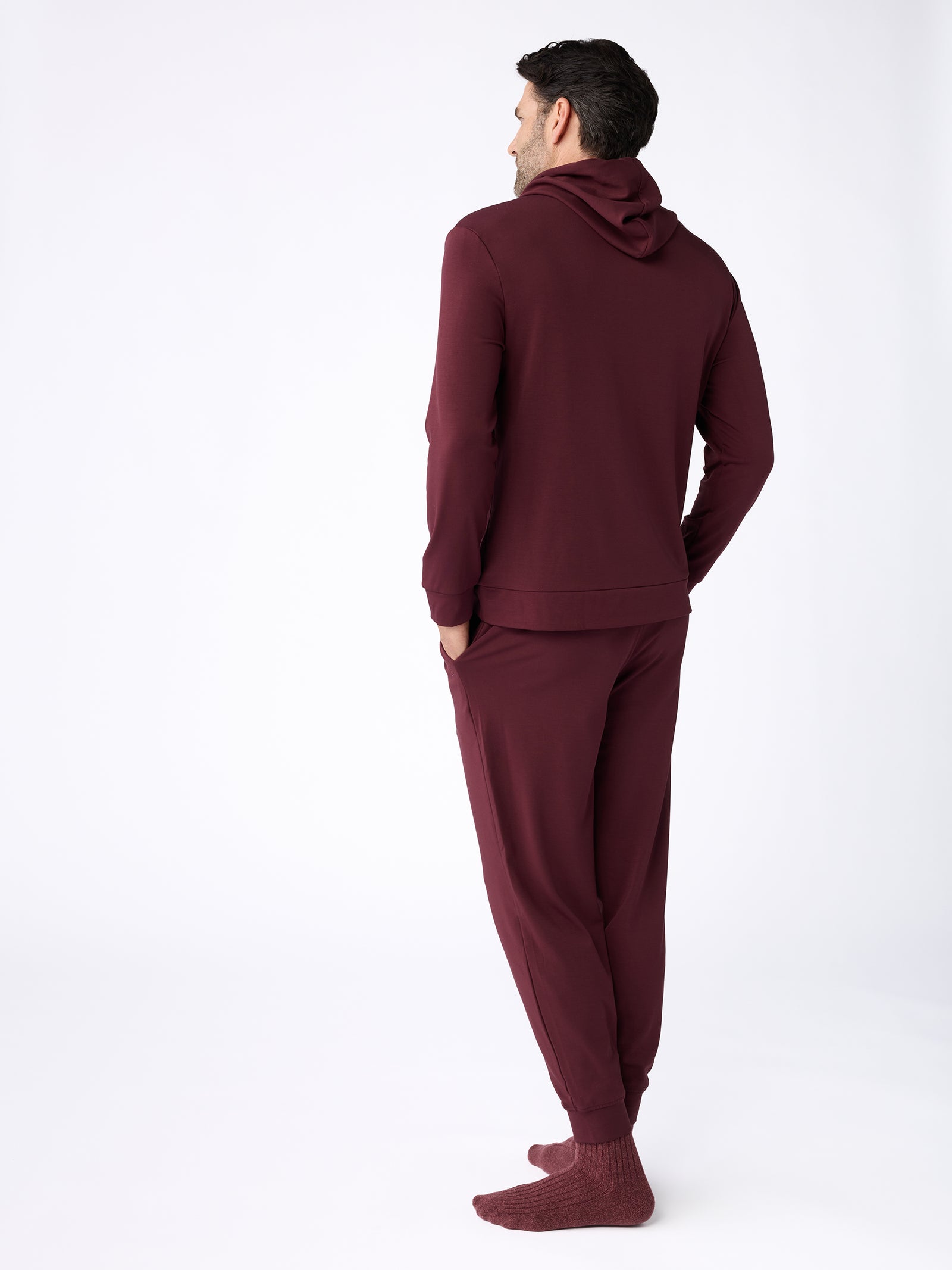 A man is facing away from the camera, dressed in a maroon hooded tracksuit with matching socks, set against a white background, while his hands rest in his pockets. He's wearing the Men's Ultra-Soft Bamboo Hoodie by Cozy Earth. 