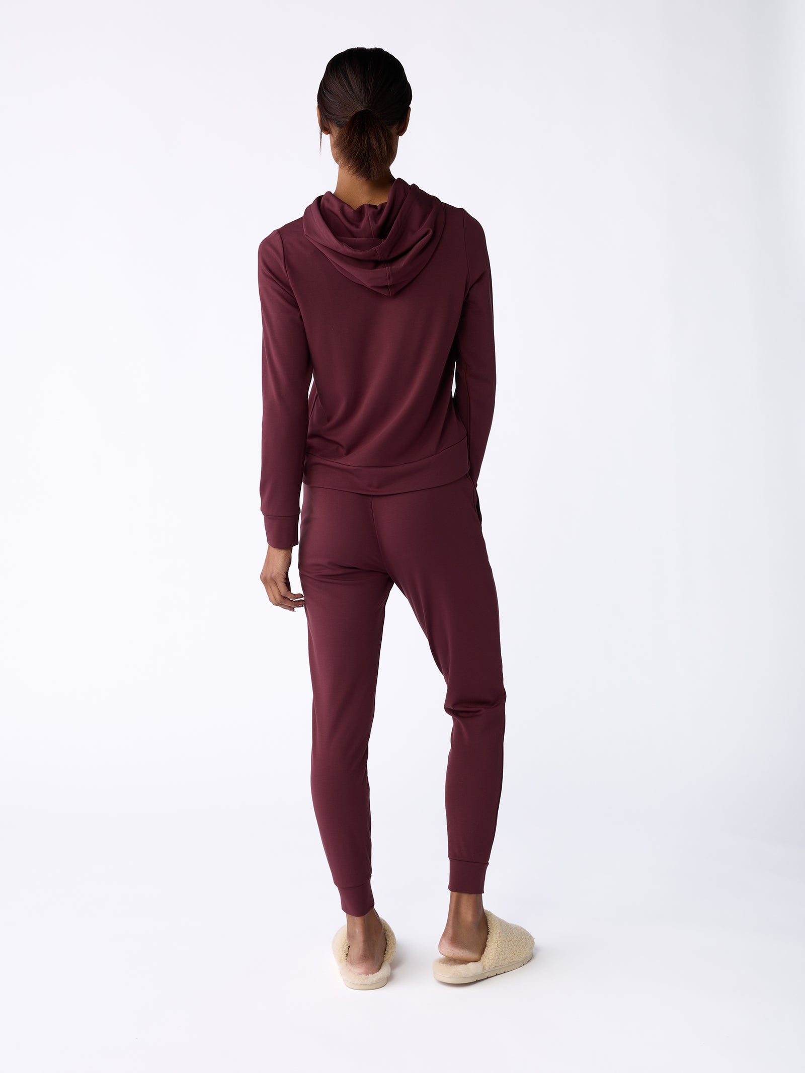 Rear view of a person wearing the Women's Ultra-Soft Bamboo Hoodie by Cozy Earth in maroon, paired with matching pants, standing against a white background. They are also wearing light-colored slippers. 