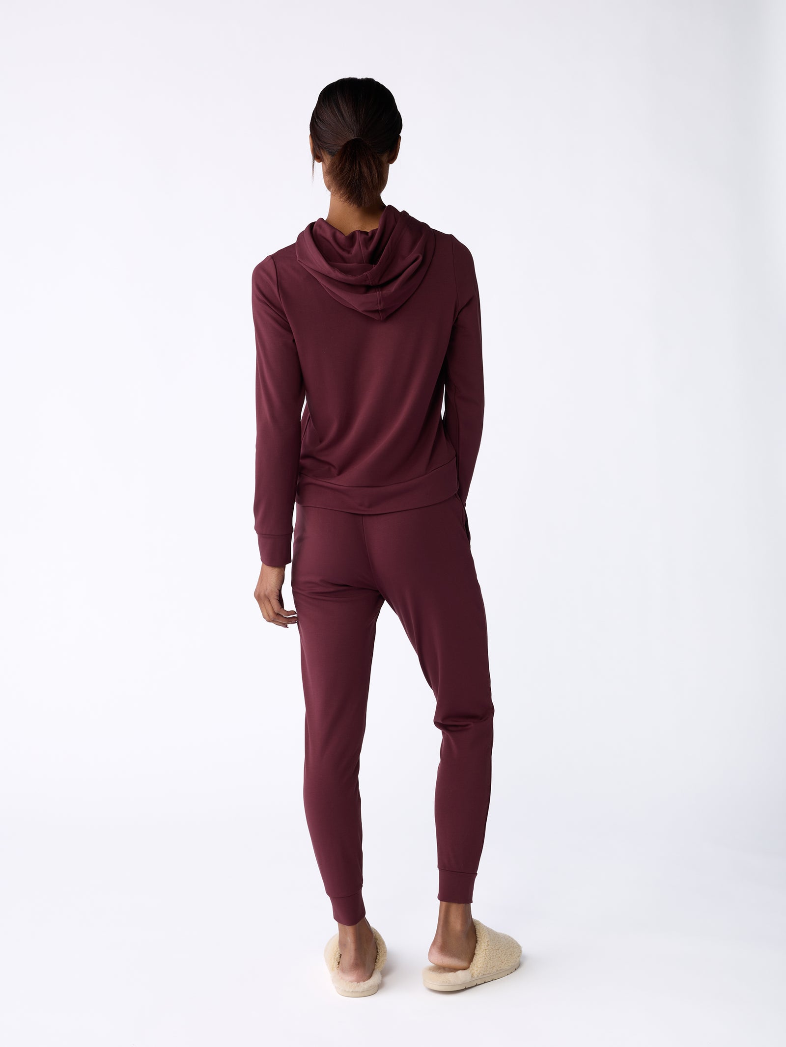 A person stands facing away, wearing Cozy Earth's Women's Bamboo Jogger Pant in maroon and beige slippers. The plain white background accentuates the outfit. 