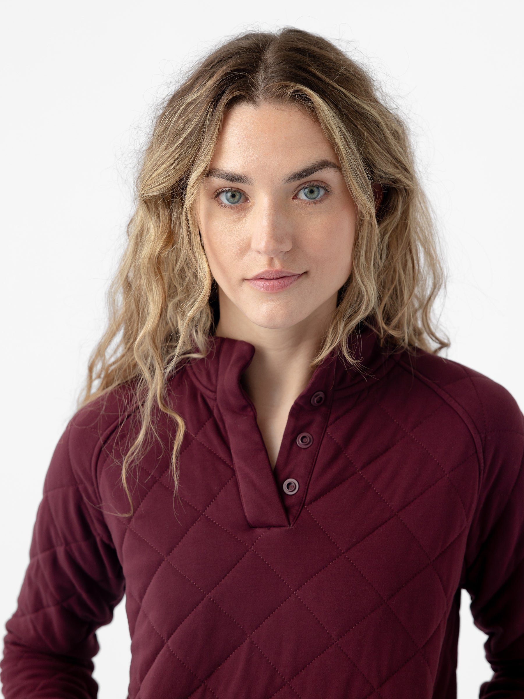A woman with long, wavy blonde hair stands facing the camera. She has blue eyes and a slight smile. She is wearing a burgundy Women's Ultra-Soft Bamboo Quilted Snap Pullover from Cozy Earth, featuring a buttoned-up high collar. The background is plain white. 