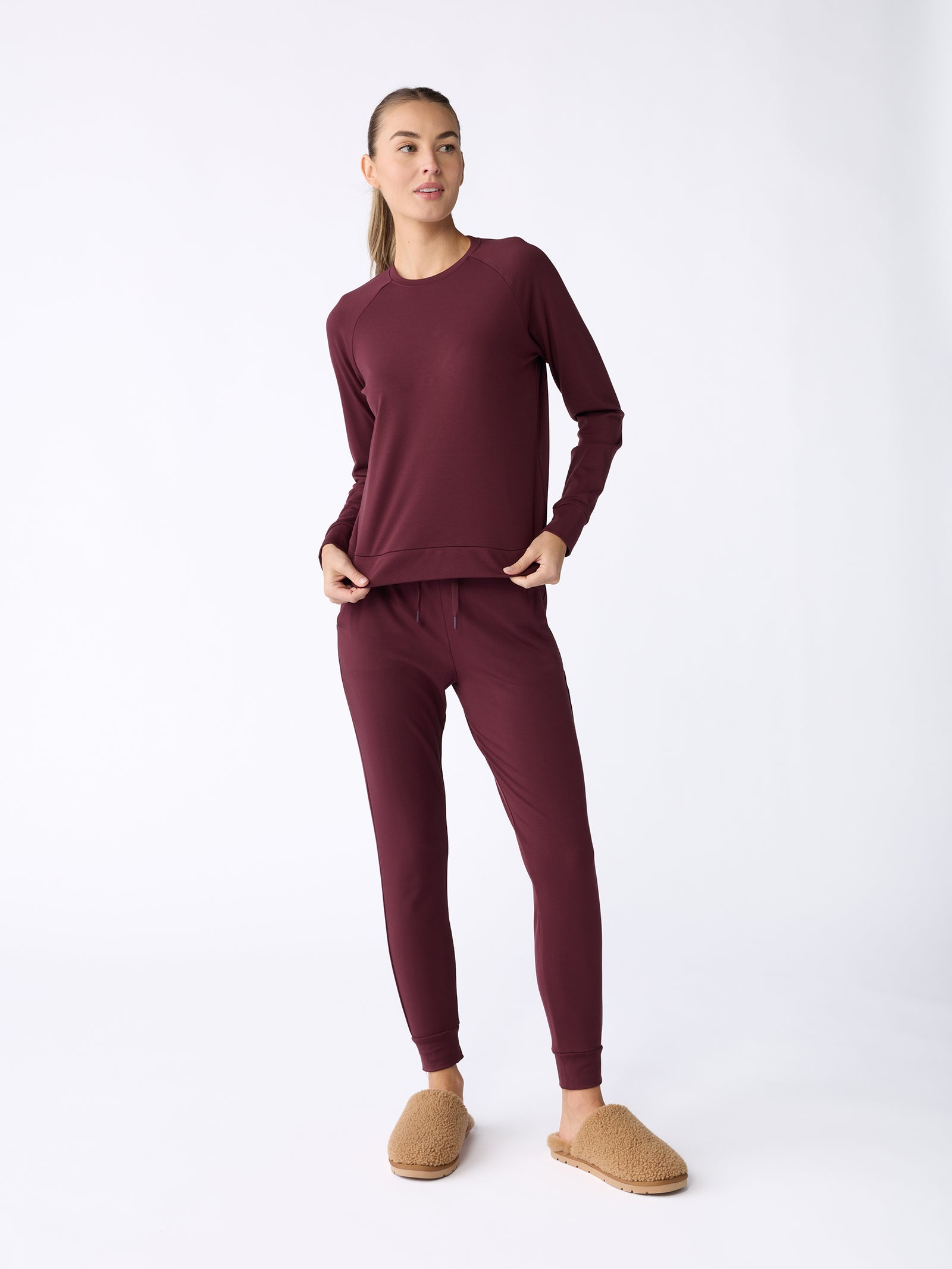 Against a plain background, a person is dressed in the Women's Ultra-Soft Bamboo Pullover Crew from Cozy Earth, paired with matching maroon jogger pants. They complete the look with tan slippers and have their hair tied back. 