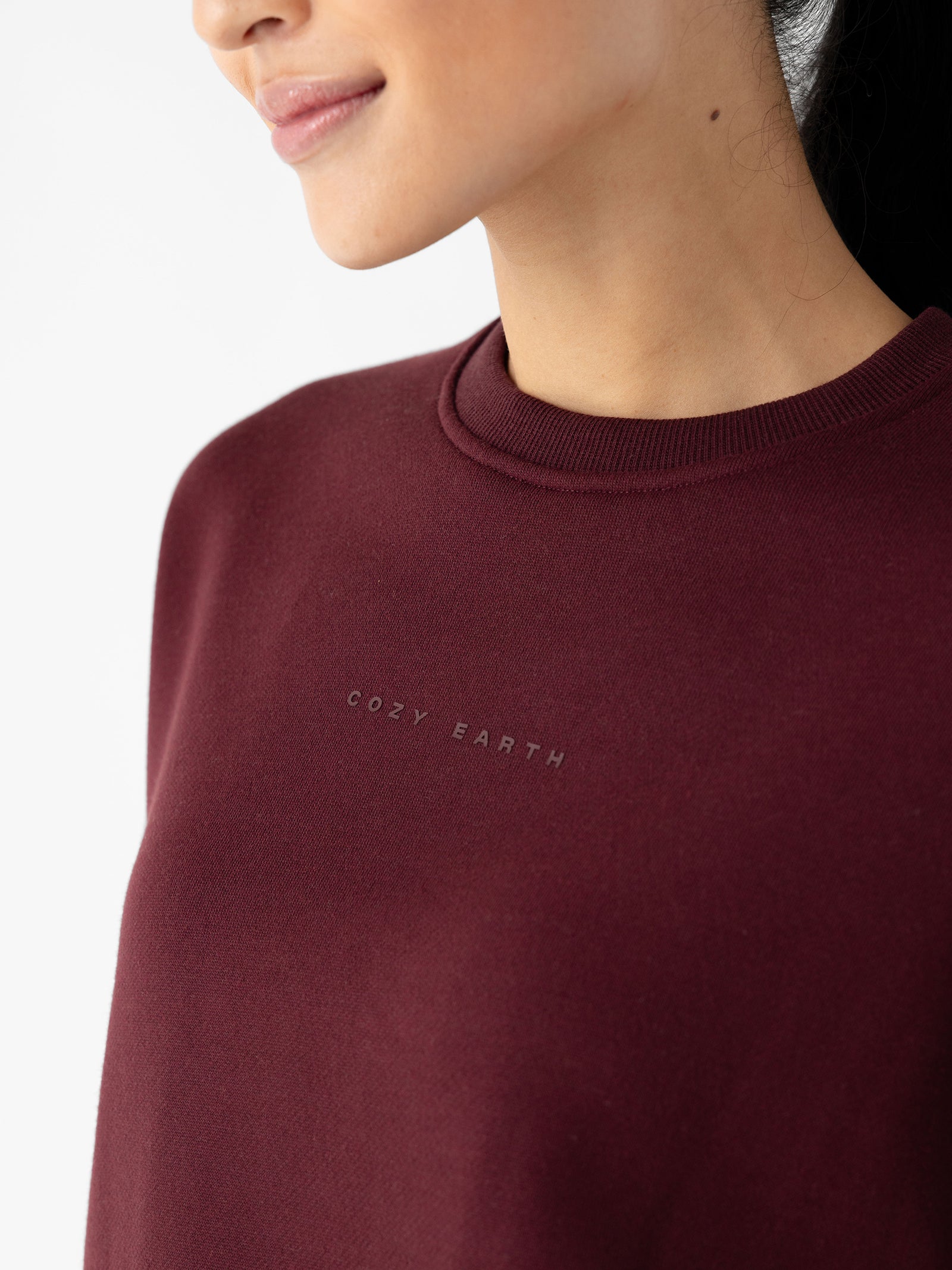 A person wearing a Women's CityScape Crewneck in deep red, with the brand name "Cozy Earth" subtly printed on the chest. The image focuses on the upper torso and part of the face against a plain white background. 