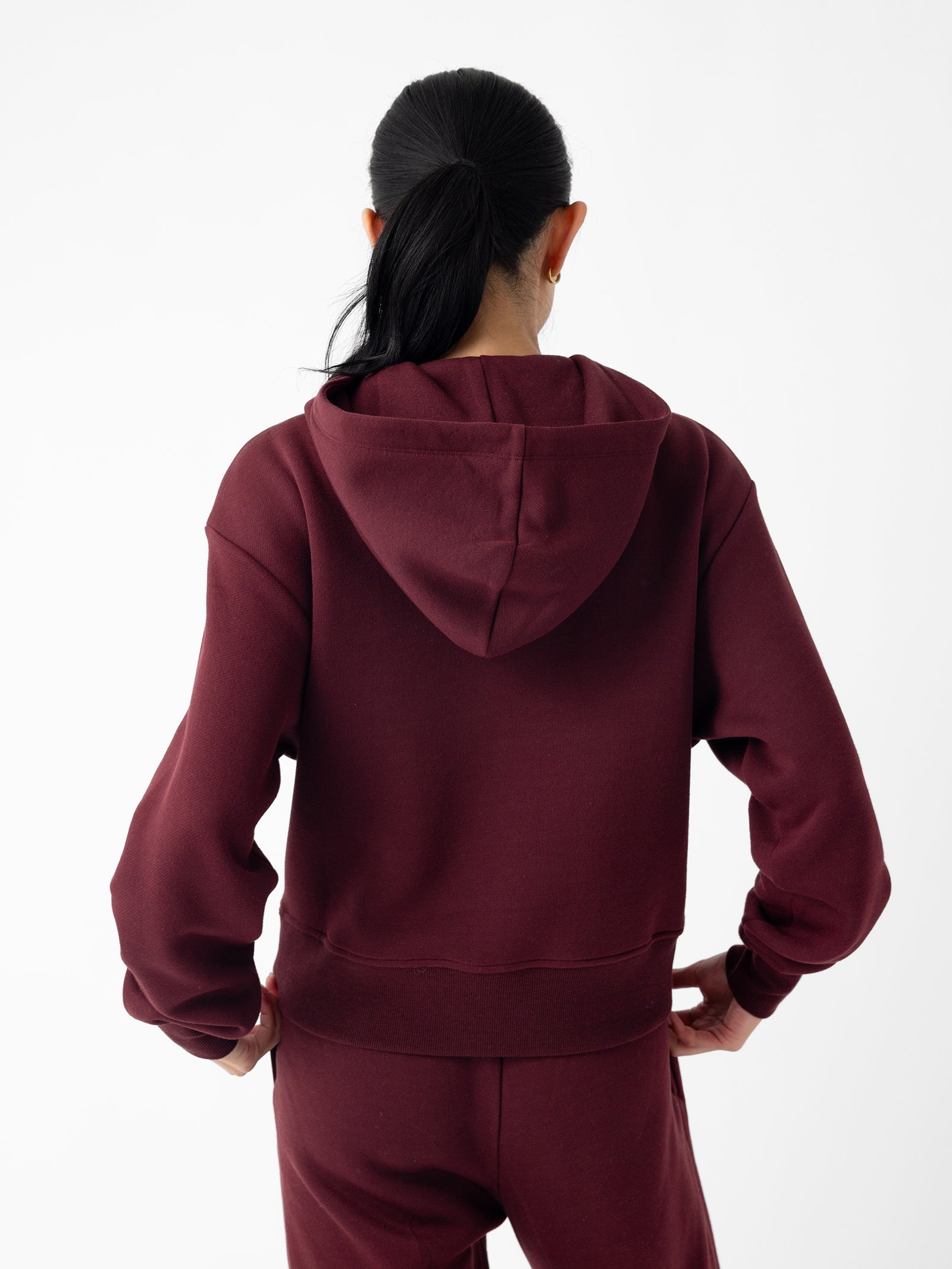 A person with long dark hair in a ponytail, wearing Cozy Earth's Women's CityScape Cropped Hoodie in burgundy and matching pants, stands with hands on hips. The person is facing away, showcasing the back of the outfit against a plain white background. 