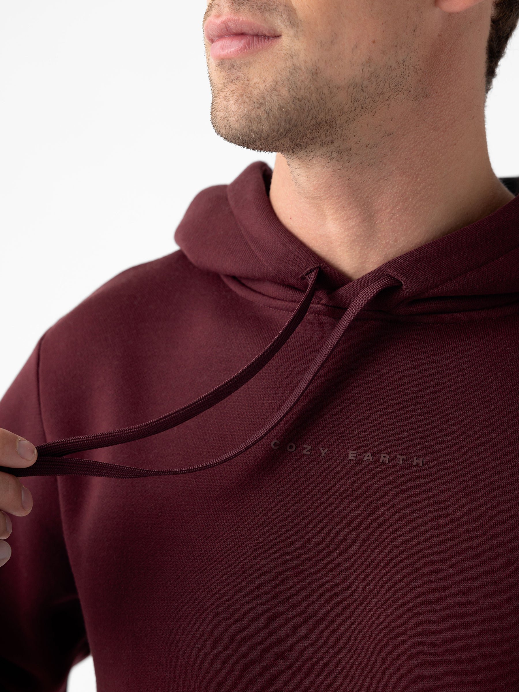 Close-up of a person wearing a maroon Men's CityScape Hoodie with the text "Cozy Earth" written on it, lightly tugging at one of the drawstrings. The face is partially out of frame, and the background is plain white. 