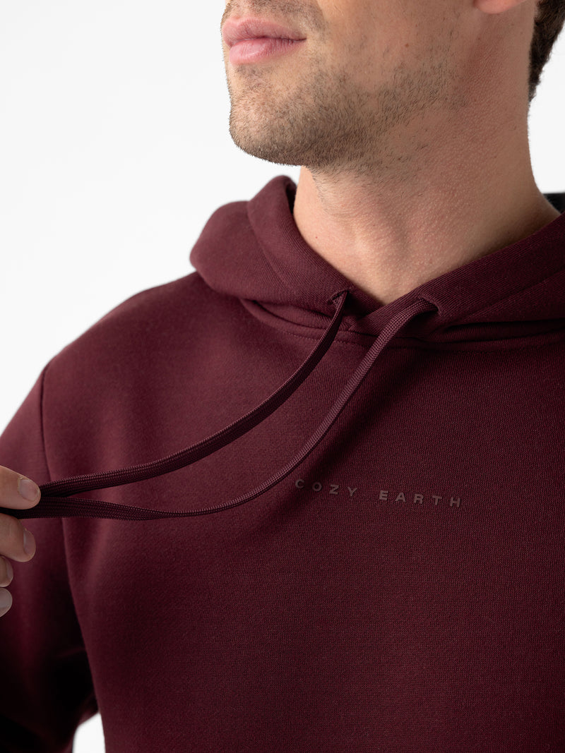 Close-up of a person wearing a maroon Men's CityScape Hoodie with the text "Cozy Earth" written on it, lightly tugging at one of the drawstrings. The face is partially out of frame, and the background is plain white.
