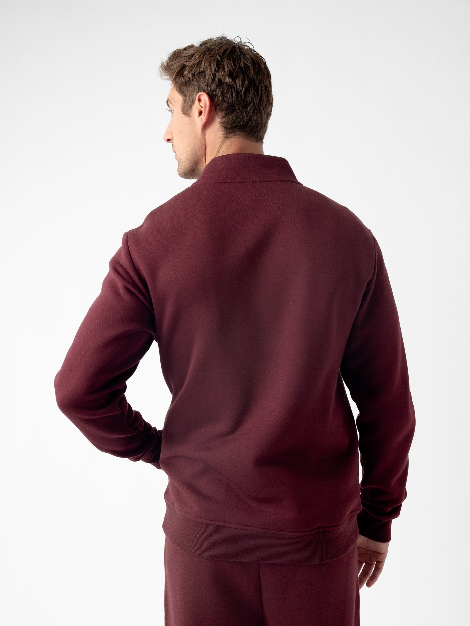 A person with short hair is standing with their back to the camera, wearing a maroon Men's CityScape Quarter Zip and matching pants by Cozy Earth. The plain white background highlights the outfit. They have one hand on their hip. 