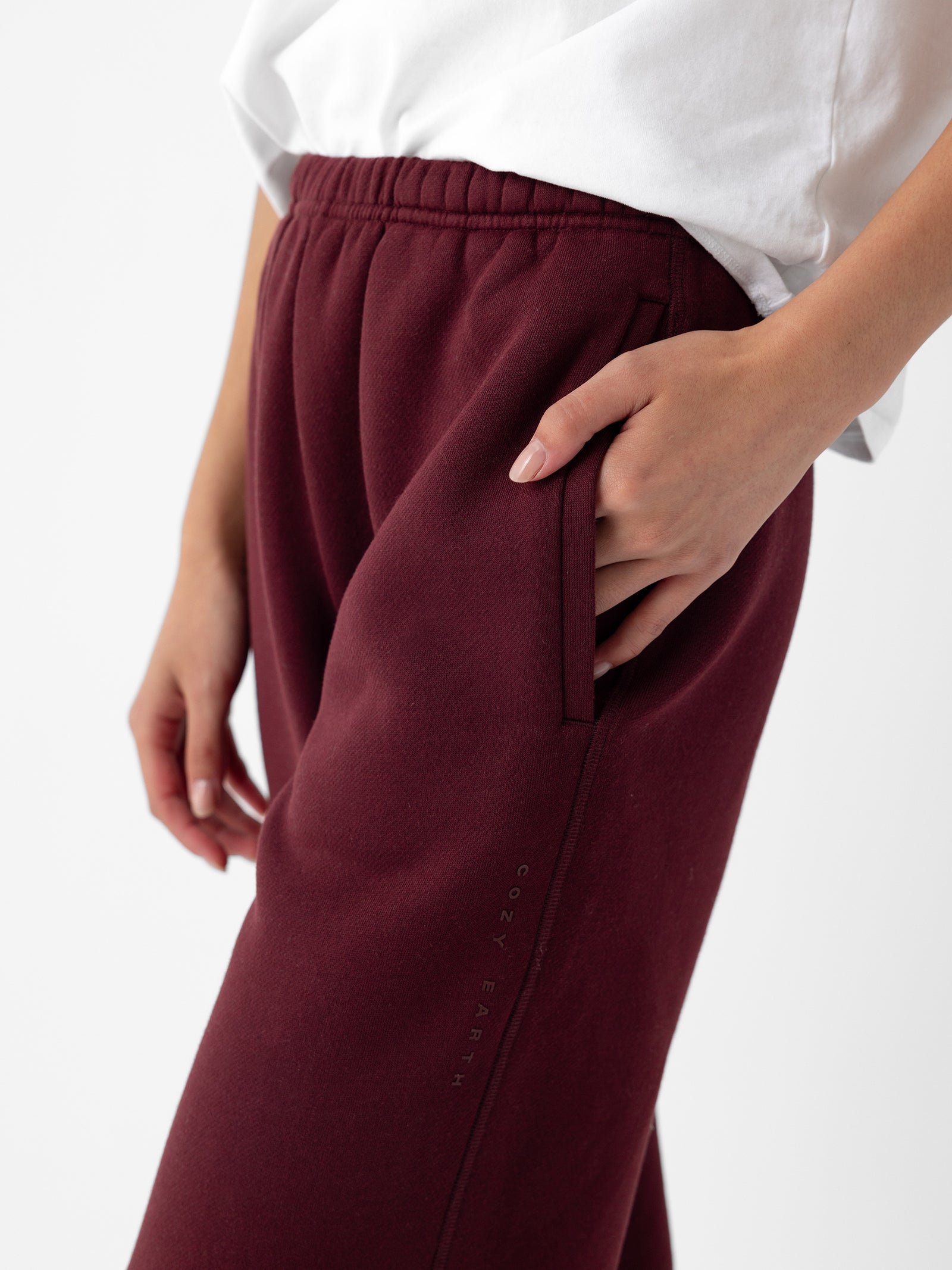 A person wearing a white shirt is shown from the waist down, with their left hand in the pocket of maroon Women's CityScape Sweatpants. The sweatpants feature the "Cozy Earth" brand name subtly printed along the left thigh. 