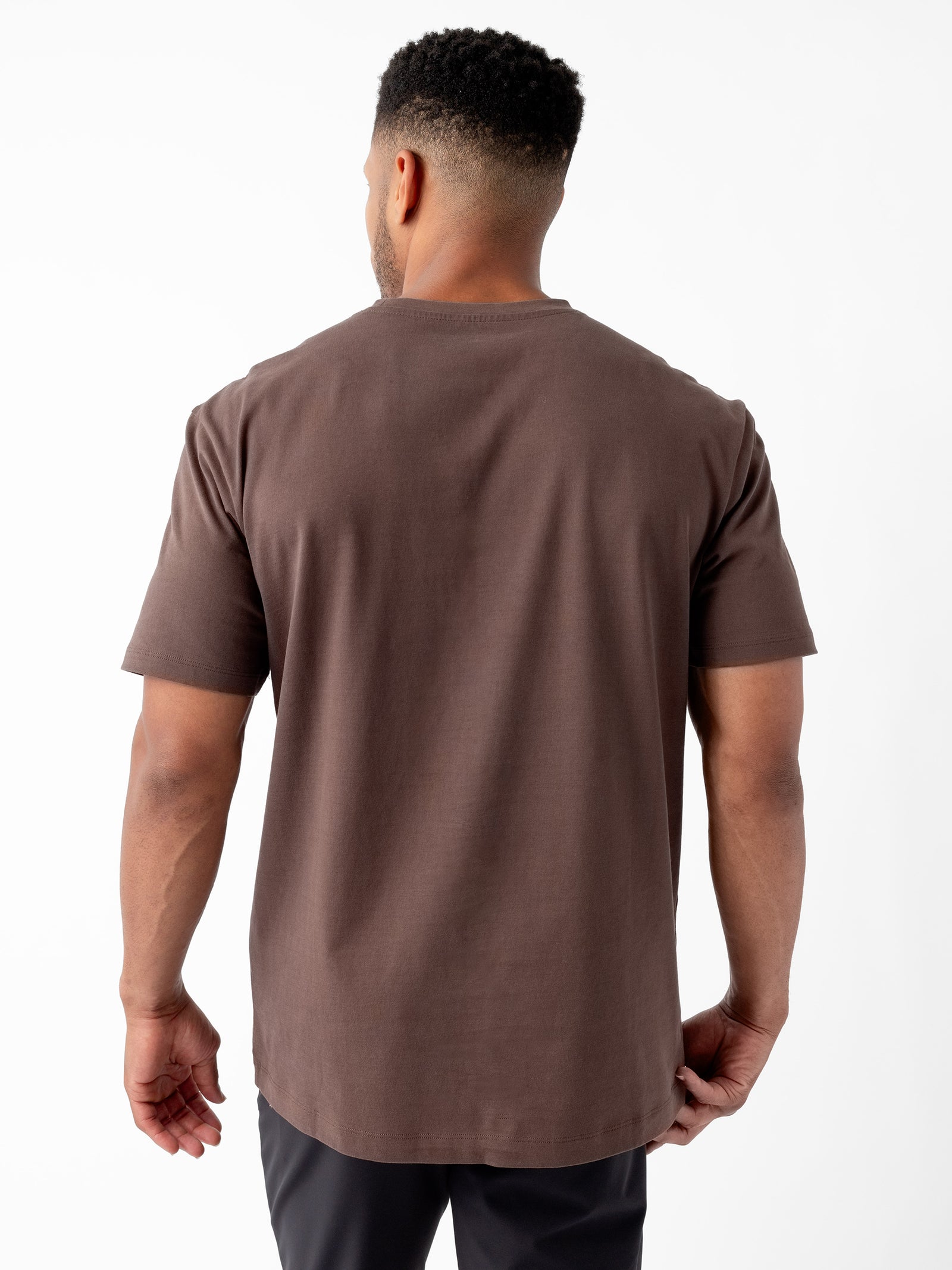 A man with short curly hair is standing with his back to the camera, wearing a plain brown Men's All Day Tee from Cozy Earth and black pants against a white background. 