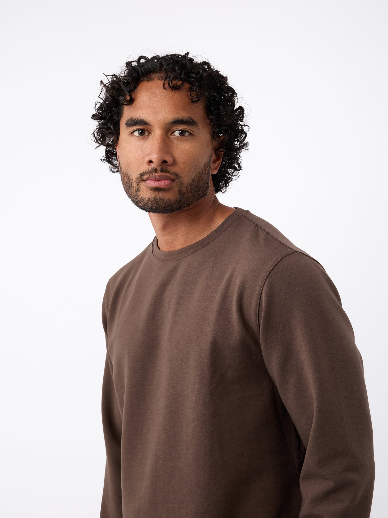 A man with curly black hair and a beard is wearing a Cozy Earth's Men's StretchTech Crewneck in brown. He is standing against a plain, light-colored background and looking directly at the camera with a neutral expression. 