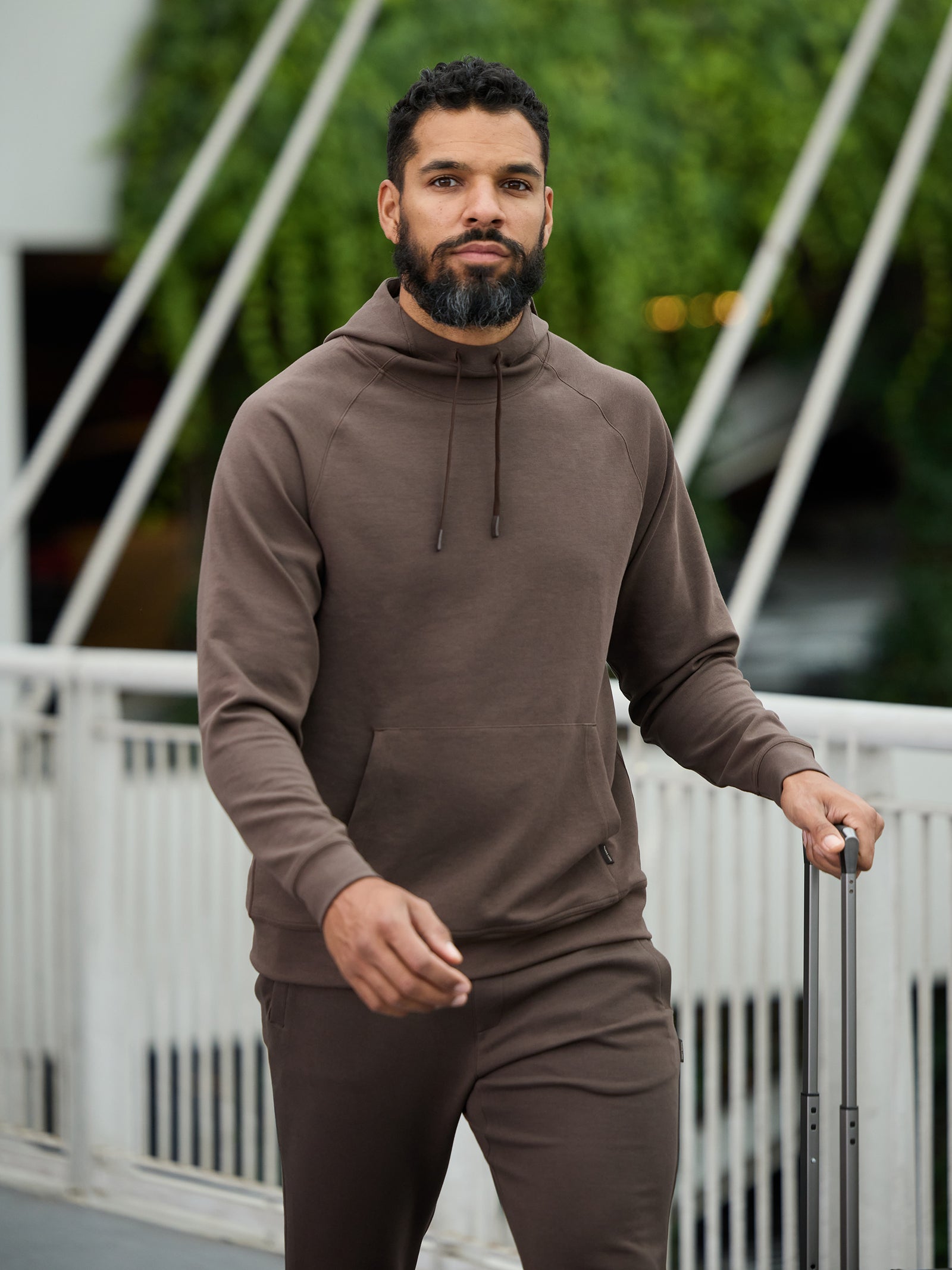 A man with a beard, dressed in a Cozy Earth Men's StretchTech Hoodie and matching pants, walks on a bridge while pulling a suitcase. A lush green backdrop is visible behind him. 