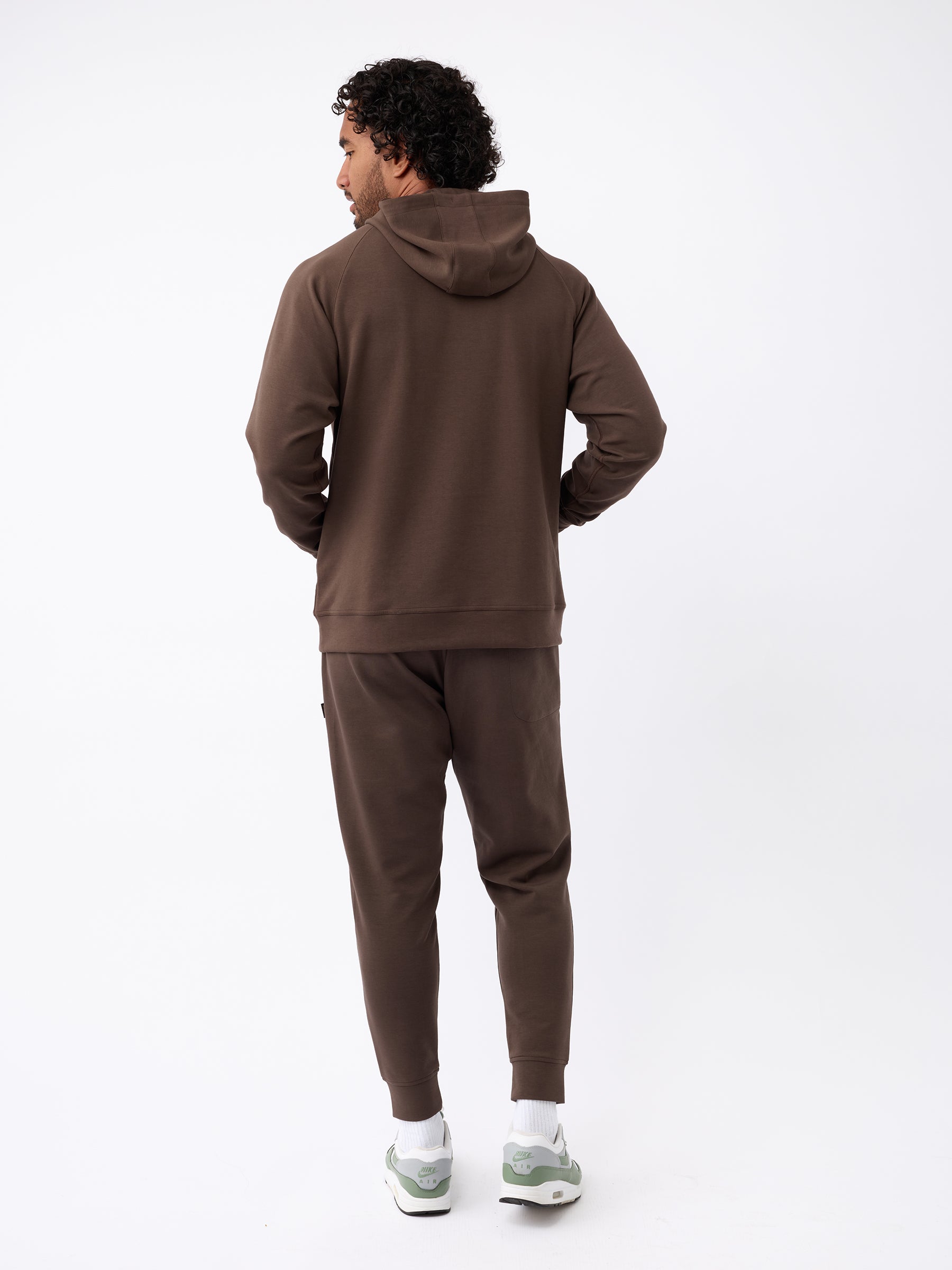 A person with curly hair stands facing away, wearing a brown Cozy Earth hoodie and matching Men's StretchTech Joggers. They have white sneakers with green accents. The background is plain white. |Color:Cacao