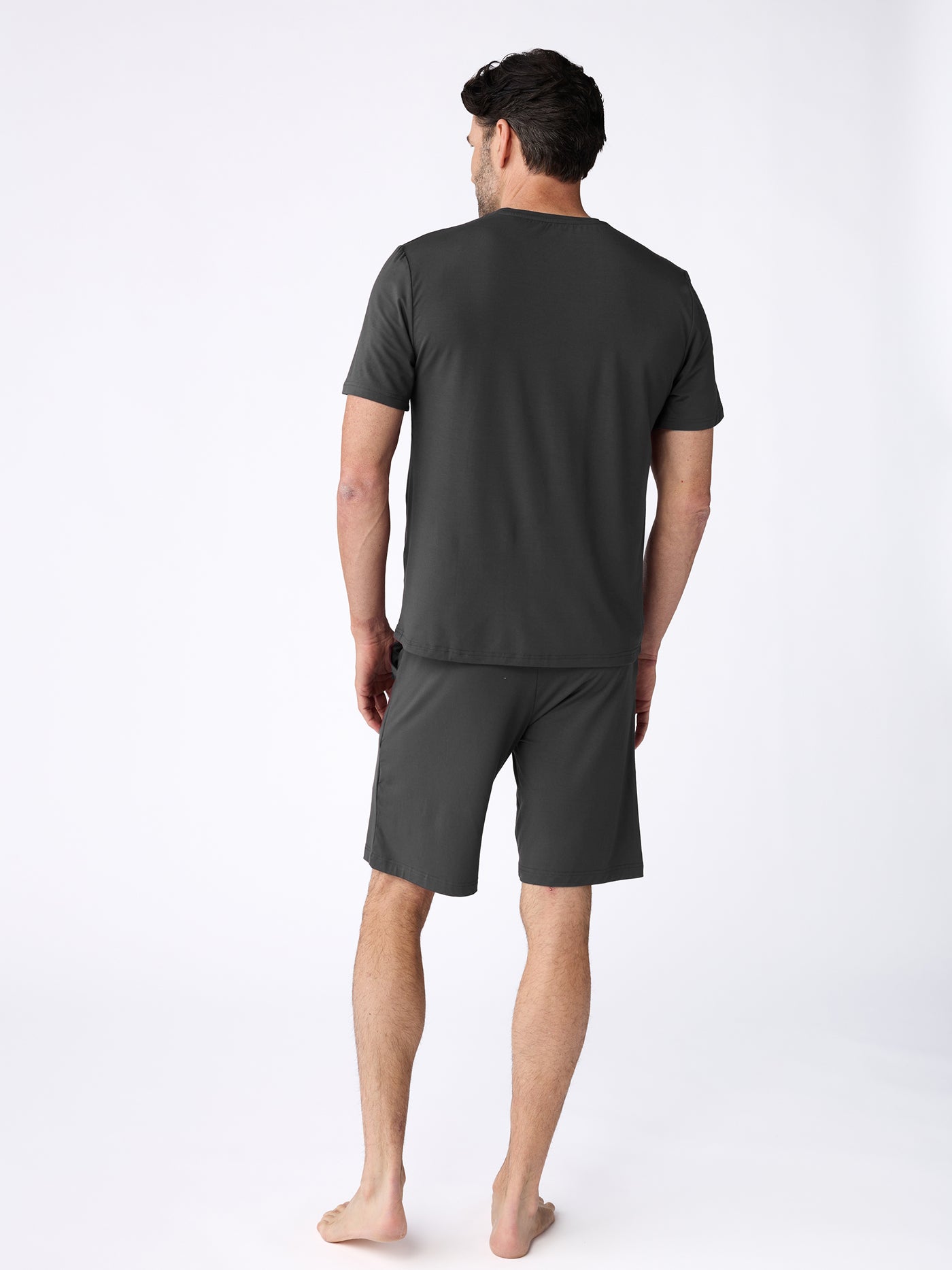 A man stands barefoot on a white background, facing away from the camera, wearing the Cozy Earth Men's Bamboo Stretch-Knit Short Sleeve Pajama Set in dark gray. |Color:Charcoal