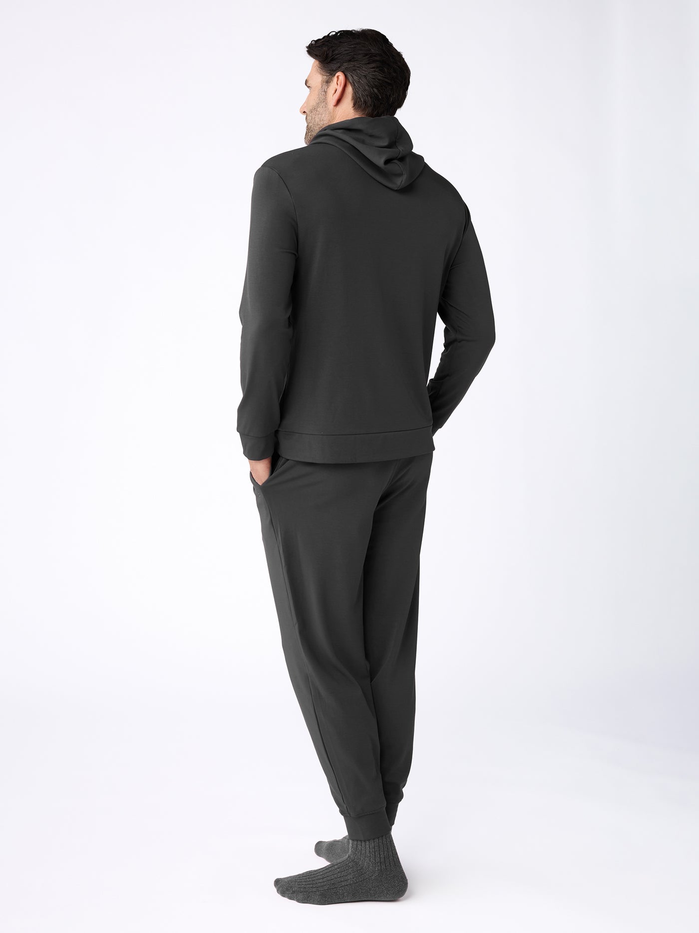 A man faces away, wearing the Cozy Earth Men's Brushed Bamboo Hoodie & Jogger Set with gray socks against a plain, light background. 