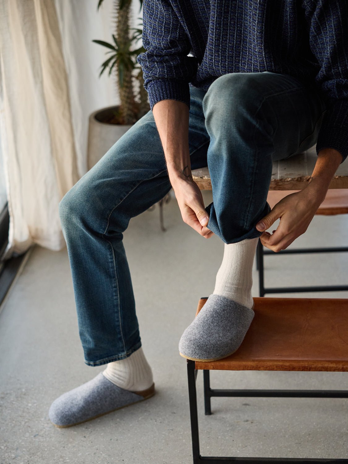 A person in a blue sweater and jeans sits on a chair, adjusting their pant leg. They're wearing white socks with gray Lakehouse Clogs by Cozy Earth. A plant and curtain are visible in the background. |Color:Charcoal