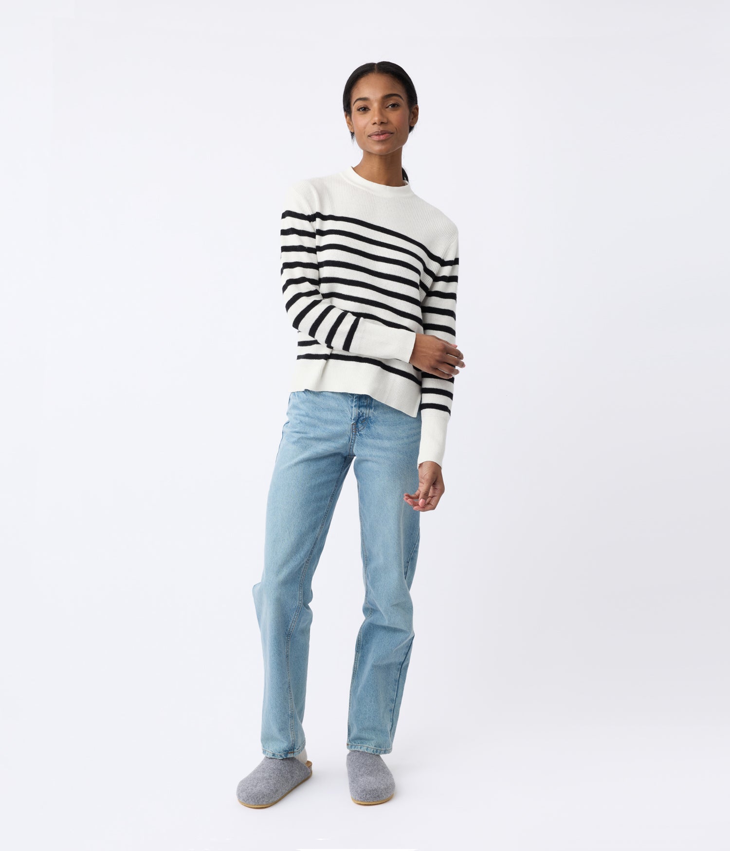 A person stands against a plain background, wearing a white sweater with black stripes, light blue jeans, and Lakehouse Clog shoes by Cozy Earth. Their pose is casual and their expression neutral. 