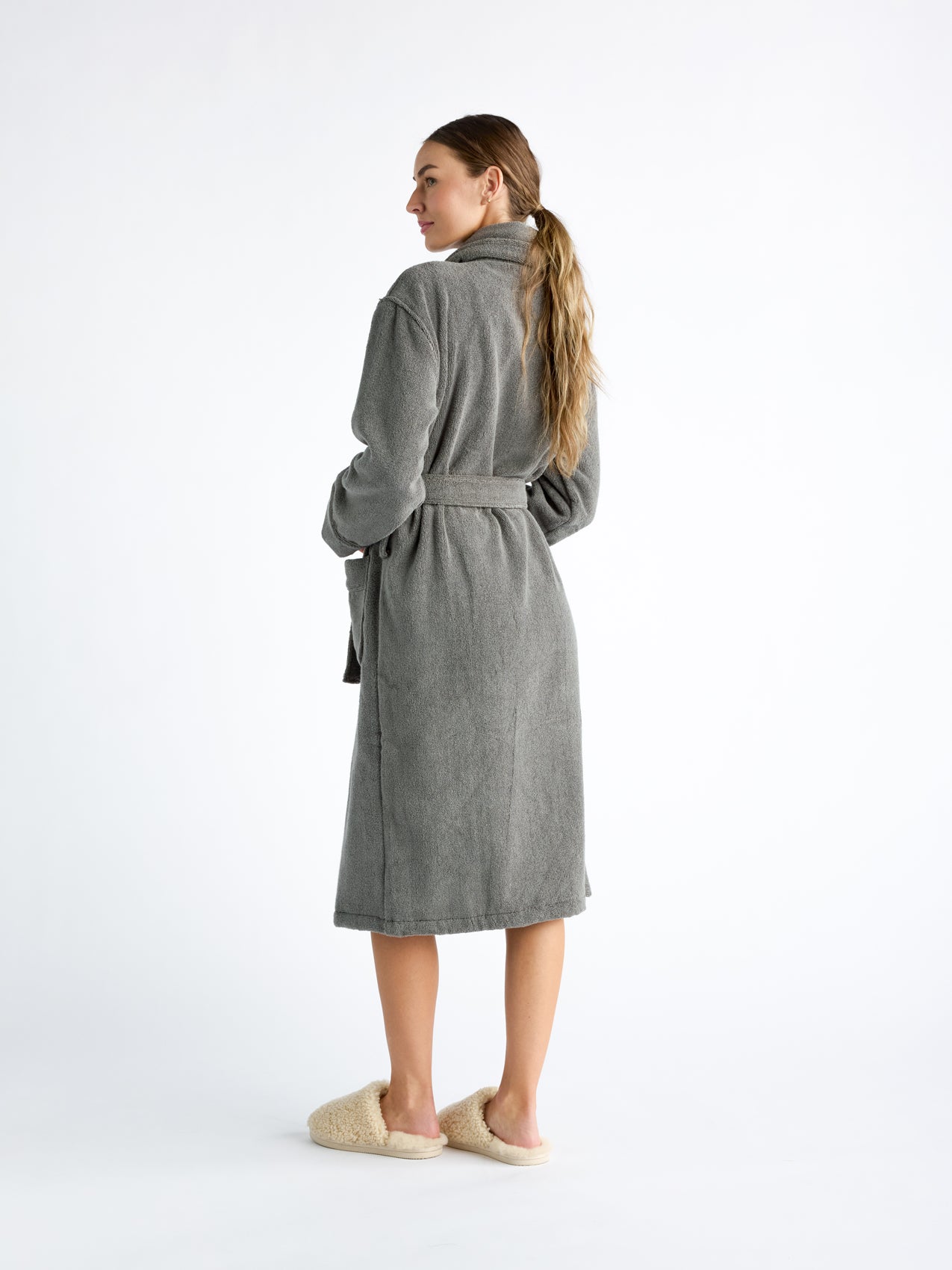 A woman is turned to the side, wearing a Luxe Bath Robe by Cozy Earth in grey. The robe is belted at the waist, and the person's long, blonde hair hangs down their back against a plain white background. 