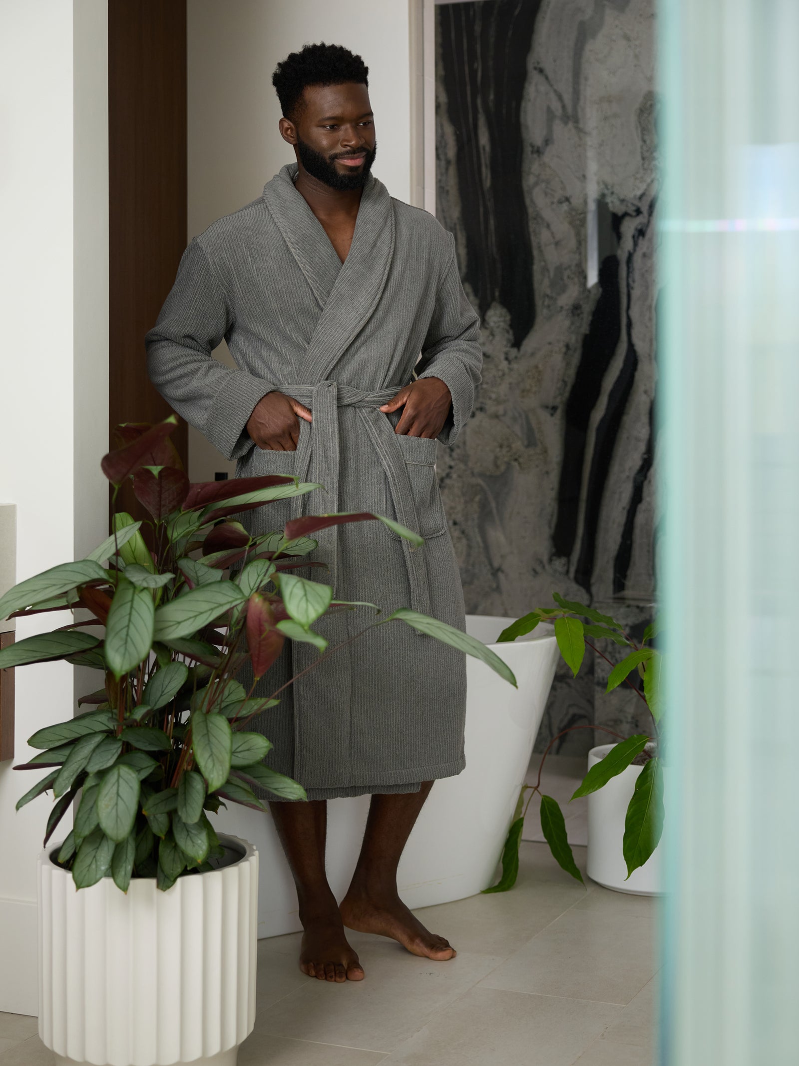 A man stands barefoot indoors, wearing a Cozy Earth Ribbed Terry Bath Robe and holding its collar with one hand. He is near a large potted plant and a marbled wall, with a soft smile on his face. The room's decor is modern and minimalistic. 