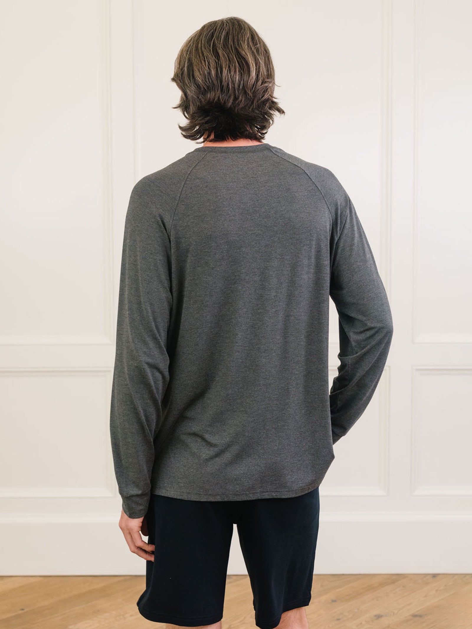 A person with shoulder-length brown hair is standing with their back to the camera, wearing a gray Men's Stretch-Knit Long Sleeve Bamboo Pajama Set from Cozy Earth and black shorts. The background features a light-colored wall with decorative molding and a wooden floor. 