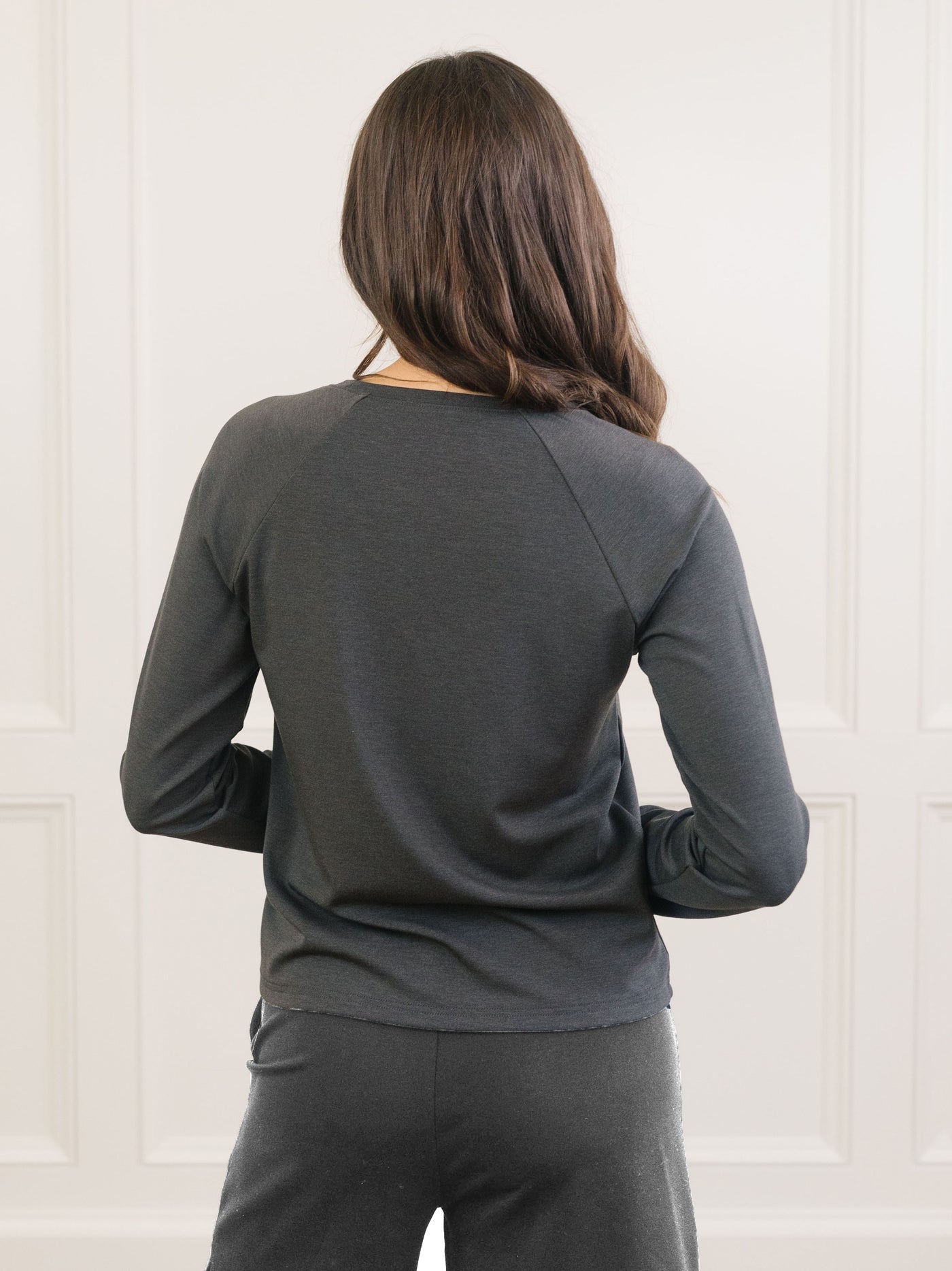 Seen from behind, a person with long brown hair is wearing the Cozy Earth Women's Ultra-Soft Bamboo Pullover Crew. They stand against a light-colored, paneled wall. 