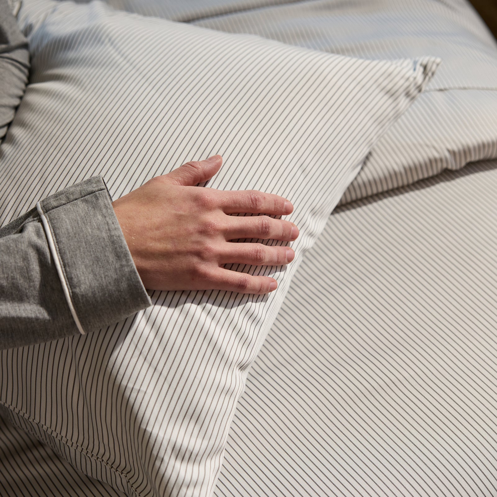 A person dressed in a gray long-sleeve top rests their hand on a Bamboo Pillowcase by Cozy Earth, placed on a matching bedspread. The setting suggests a cozy, peaceful environment. 