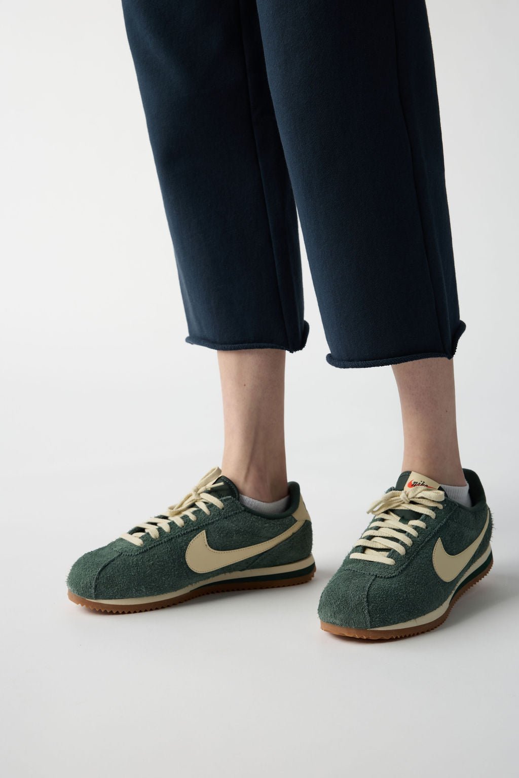 A person is dressed in Cozy Earth's Women's Chelsea Set, featuring dark green sneakers with a beige swoosh and gum sole, paired with dark blue cropped pants against a light background. 