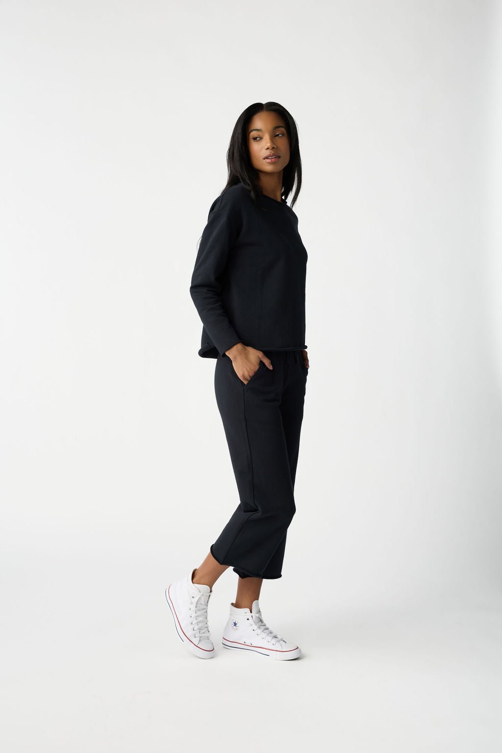 Wearing Cozy Earth's Women's Chelsea Set, featuring a dark long-sleeve top and matching cropped pants, a person stands against a white backdrop in white high-top sneakers, with one hand in their pocket and looking at the camera. 