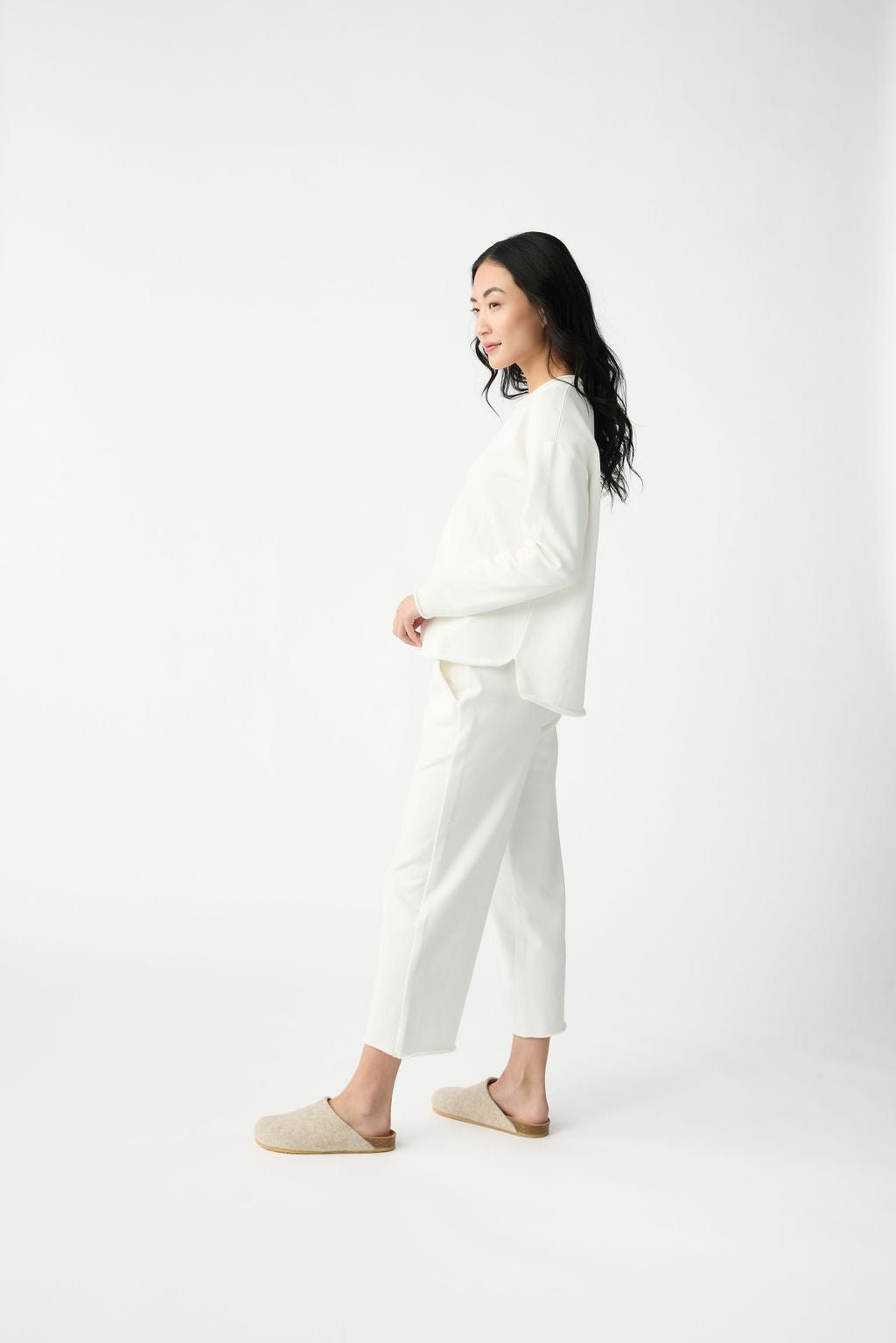 In a bright, minimalist room, a person with long dark hair stands sideways wearing the Women's Chelsea Set by Cozy Earth—an all-white ensemble featuring a loose top and cropped pants, paired with beige slip-on shoes against a plain white background. |Color:Bone