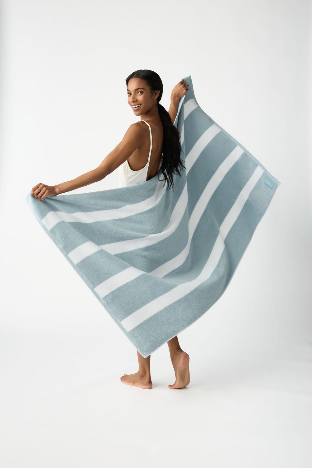 A person with long dark hair smiles, barefoot and dressed in a white outfit, while holding the Cozy Earth Classic Stripe Resort Towel wrapped around them against a plain white backdrop. 