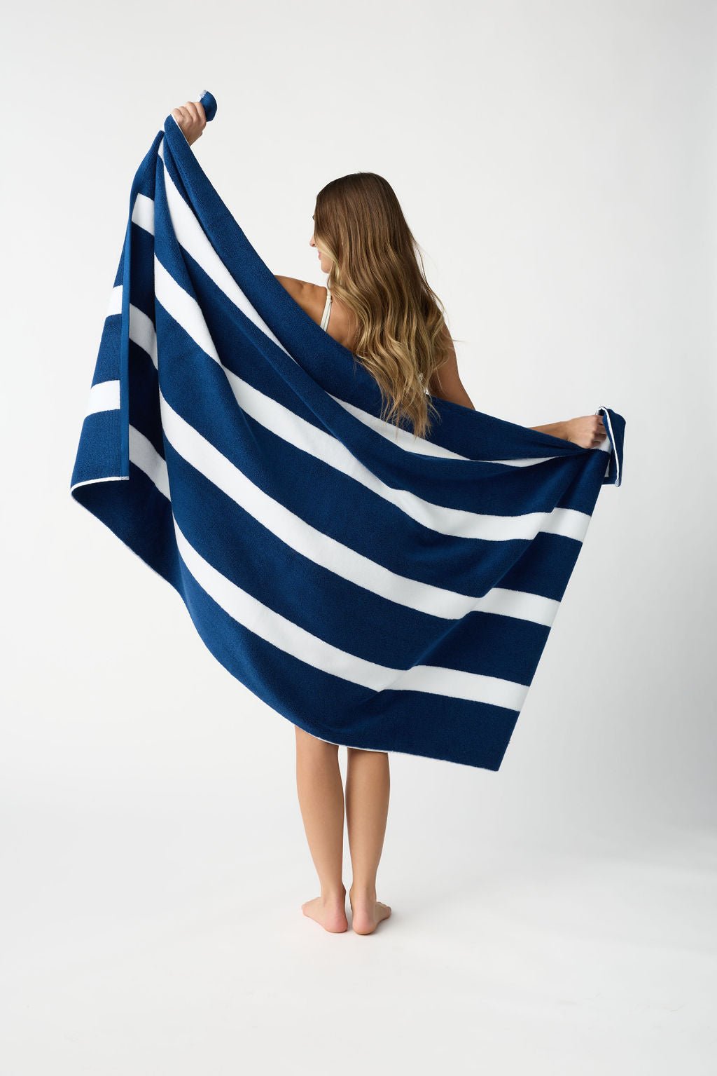 A person with long hair stands barefoot on a plain background, holding out the Classic Stripe Resort Towel from Cozy Earth. The large blue and white striped towel drapes around them as they face away from the camera. 