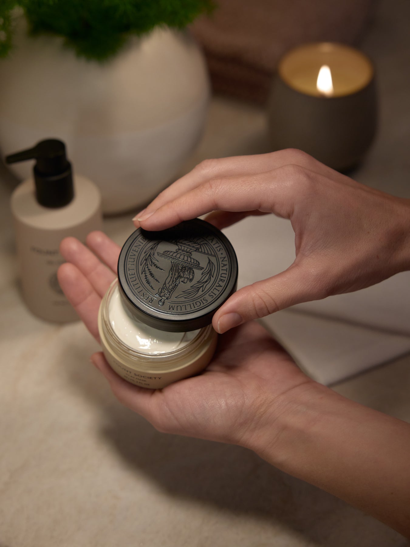 Hands opening a jar of Cozy Earth's Clay Mask, highlighting the product's texture. A pump bottle, towel, potted plant, and lit candle blur in the background to create a serene, spa-like ambiance. 