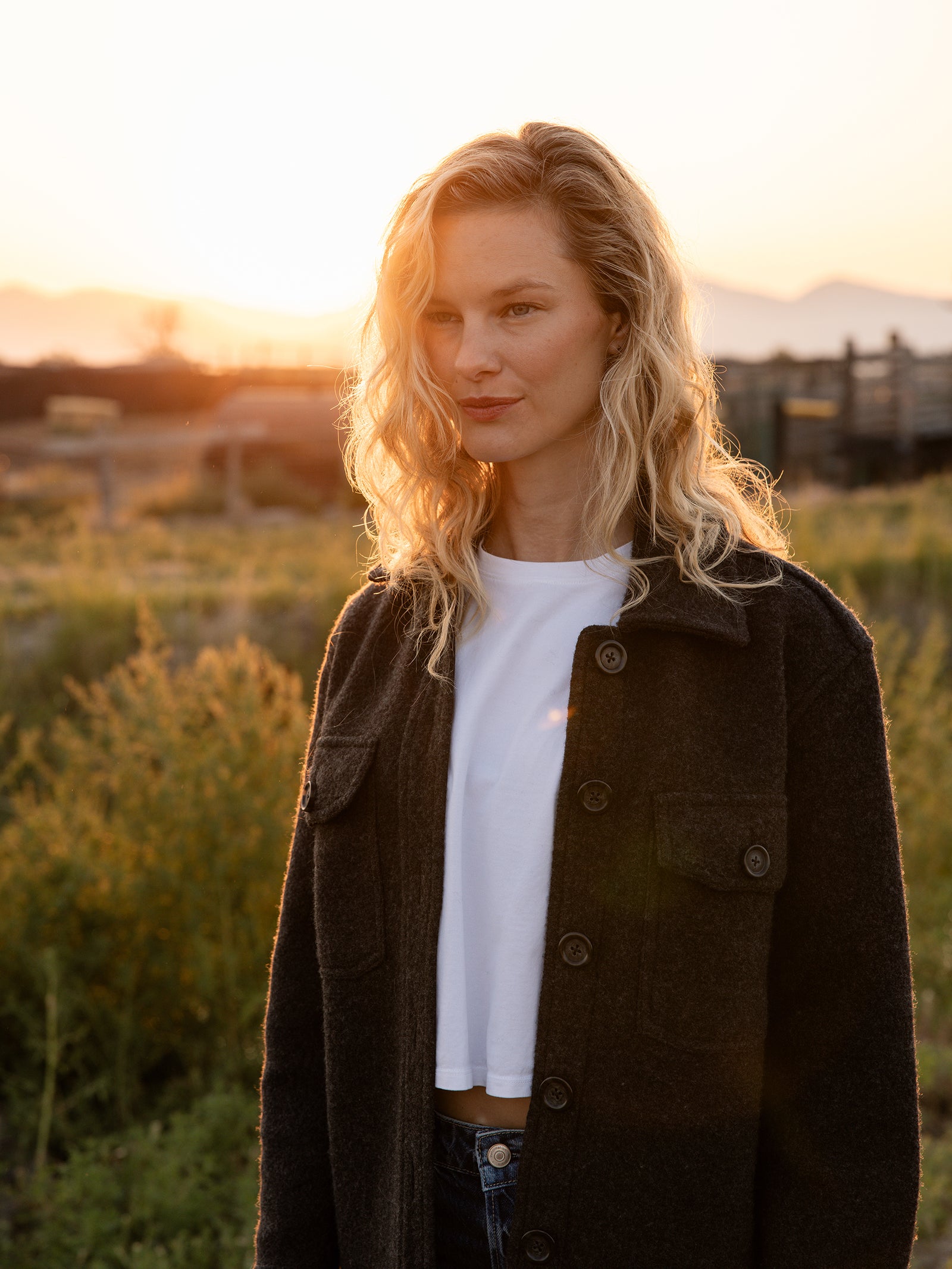 A woman with wavy blonde hair stands outdoors at sunset, wearing a dark coat over a white shirt. She is looking slightly to the side. The background features fields, some plants, and distant mountains bathed in the warm light of the setting sun. She is elegantly dressed in a Cozy Earth Women's Boucle Shacket. 