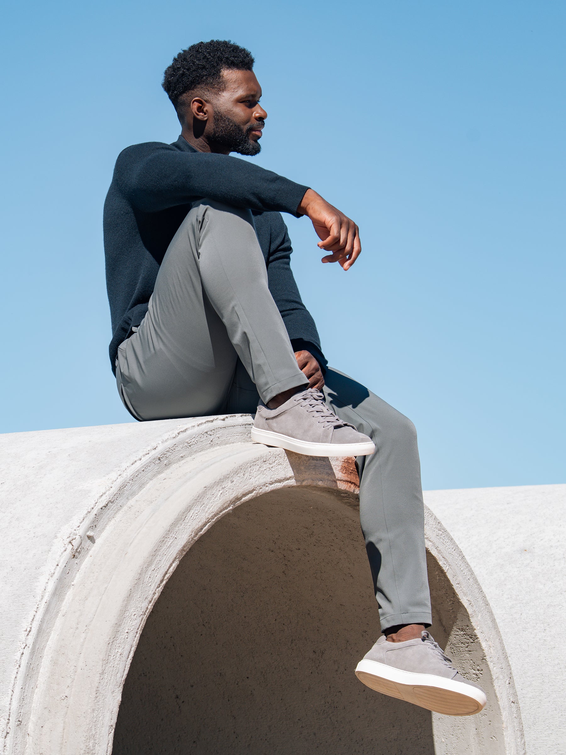 A man dressed in a dark sweater and Cozy Earth's Men's Everywhere Pant 30L in light gray sits atop a large concrete pipe against a clear blue sky. He is looking to the side with one arm resting on his knee and the other hanging down, wearing gray sneakers with white soles. |Color:Coal