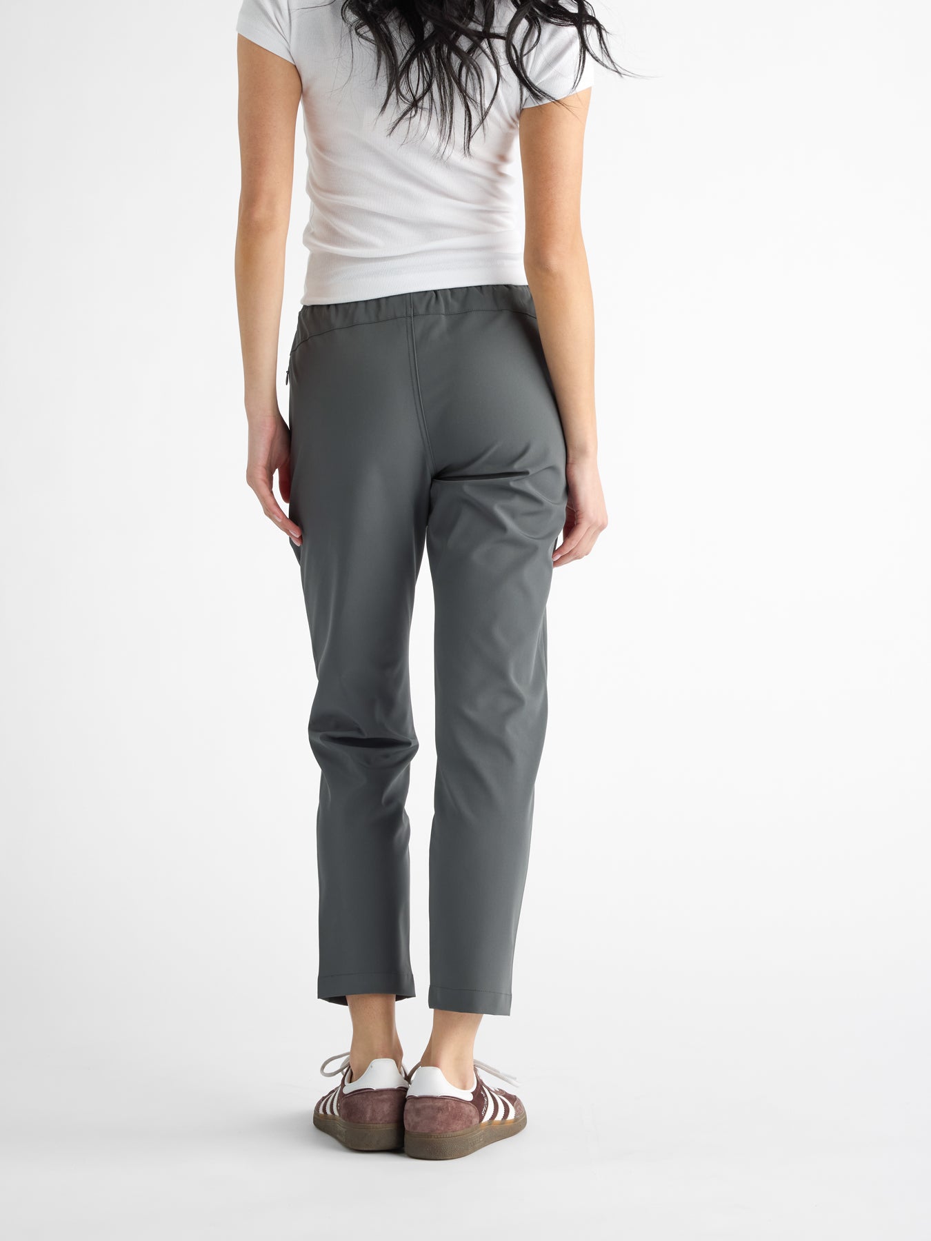 Rear view of a person with long hair wearing a white t-shirt and Cozy Earth's Women's Always Cropped Pant in gray. They are standing on a white background, sporting brown shoes with white stripes, though their head isn't visible. 