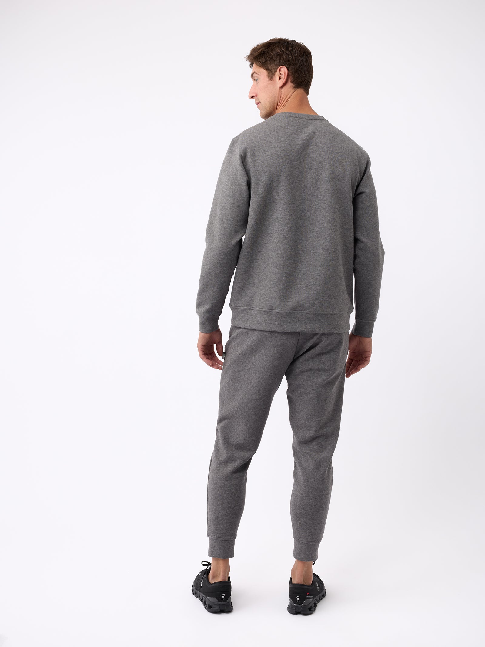 Dressed in a Men's StretchTech Crewneck sweater from Cozy Earth and matching joggers, a person stands facing away while sporting black sneakers against a plain white background. 