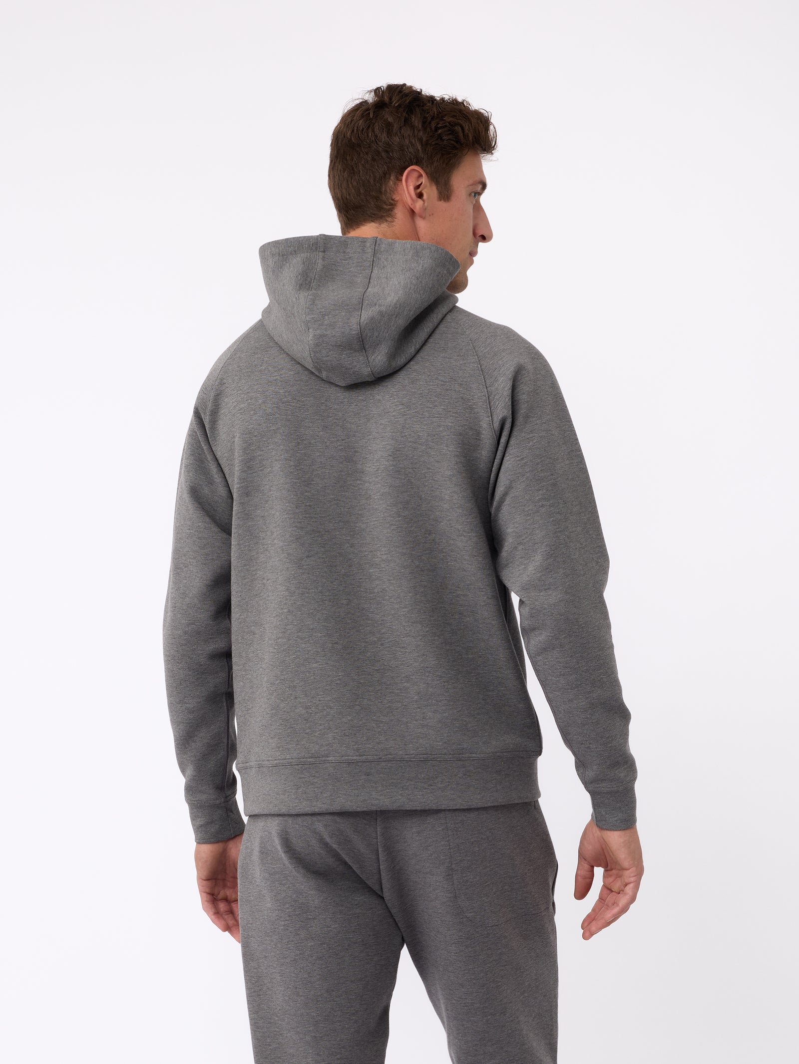 A person wearing a Cozy Earth Men's StretchTech Hoodie and matching gray sweatpants stands with their back to the camera. The image emphasizes the hood and relaxed fit of the clothing against a plain white background. 