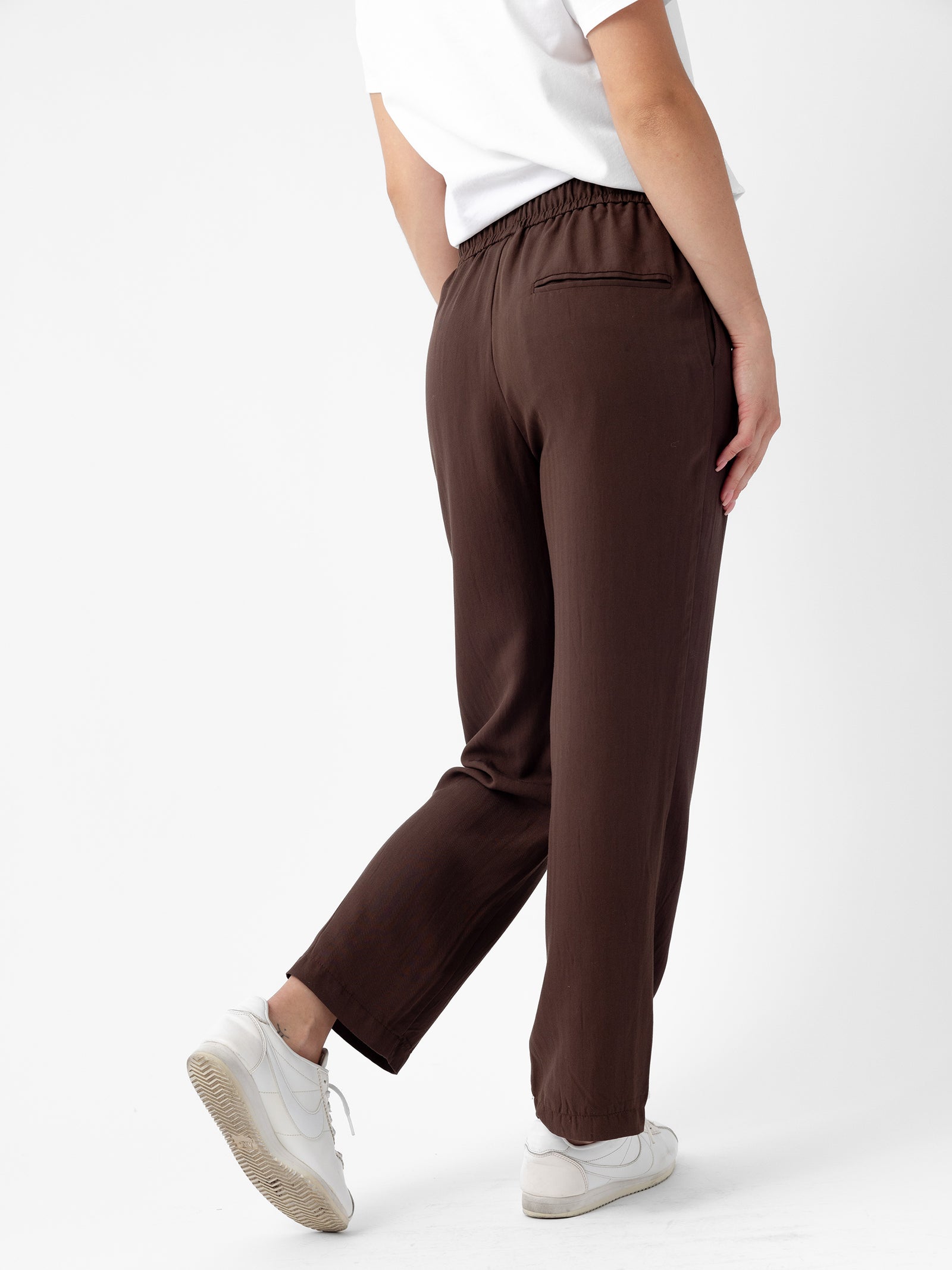 A person is shown from the back, wearing a white top and Cozy Earth's Women's Sunset Cropped Pant in brown. The left leg of the person is slightly bent and they are wearing white sneakers. The background is plain white. 