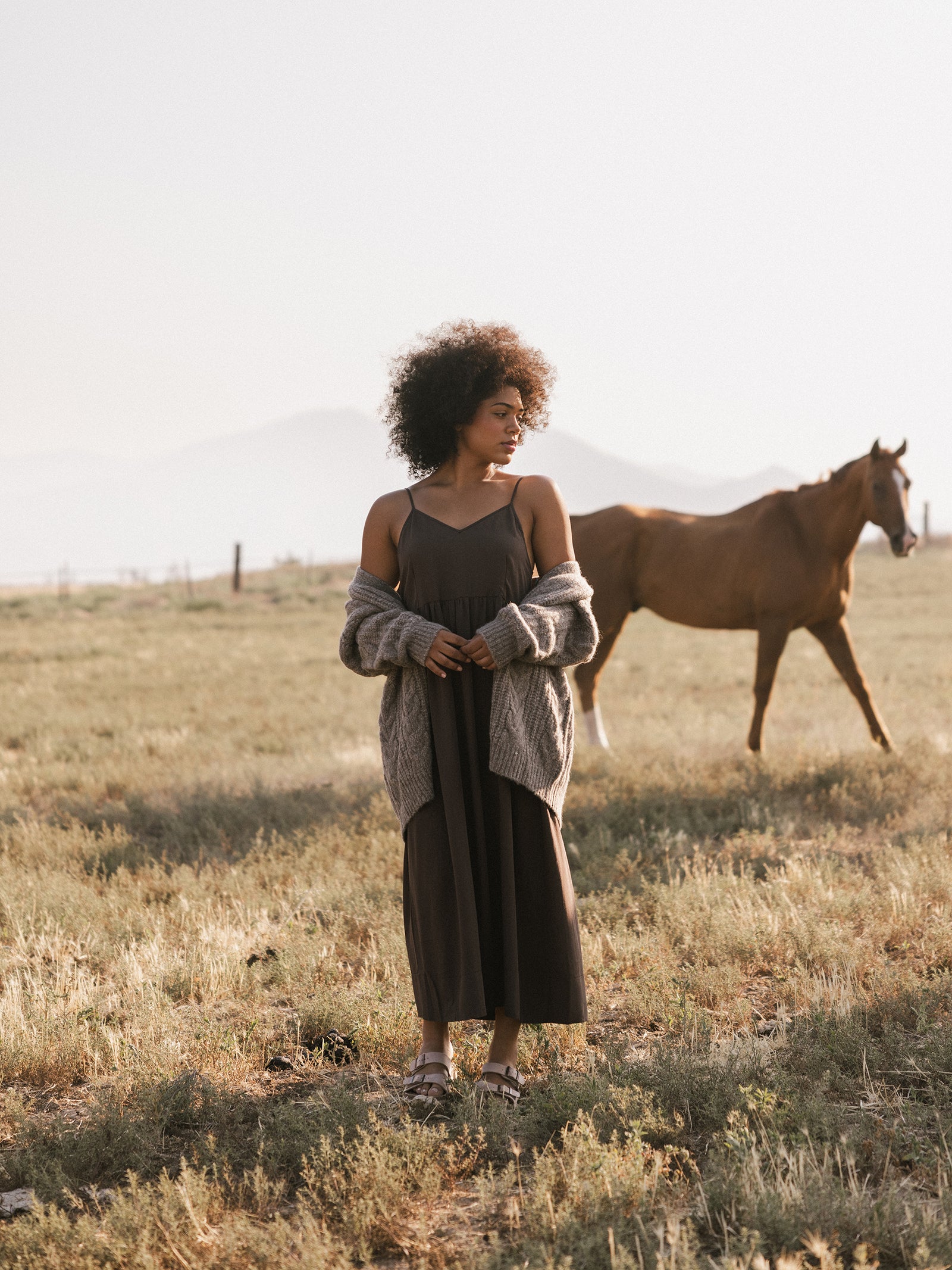 A person with curly hair stands in a grassy field wearing Cozy Earth's Women's Sunset Gathered Dress and a light gray cardigan, with eyes gazing downward. In the background, a brown horse walks casually. Misty mountains are visible in the distance under a pale sky. 