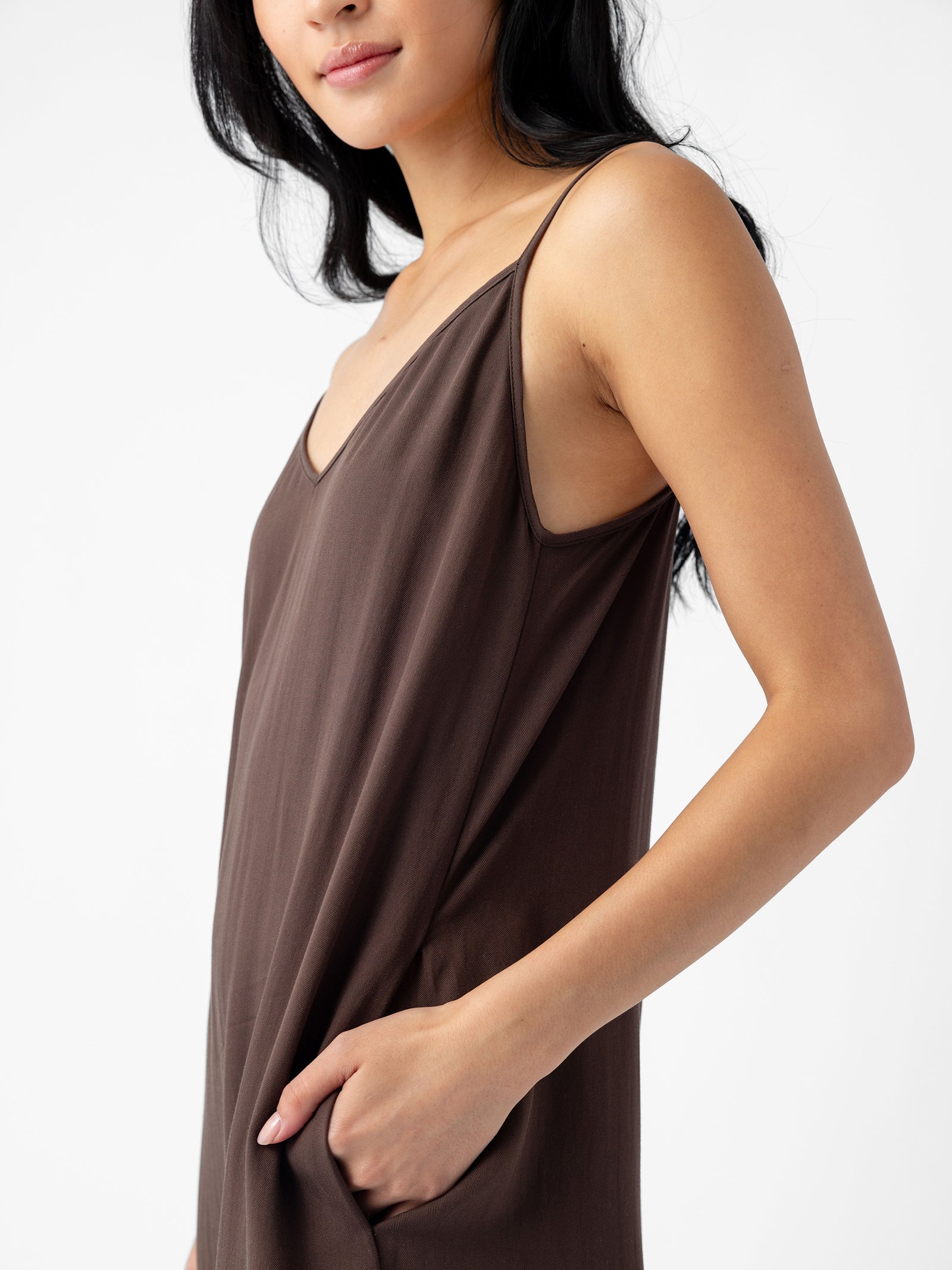 A person with long dark hair wears a Cozy Earth Women's Sunset Slip Dress, featuring a sleeveless design and dark brown color with a V-neckline. Their right hand is casually placed in the dress pocket, and they are slightly turned to one side against a plain white background. 