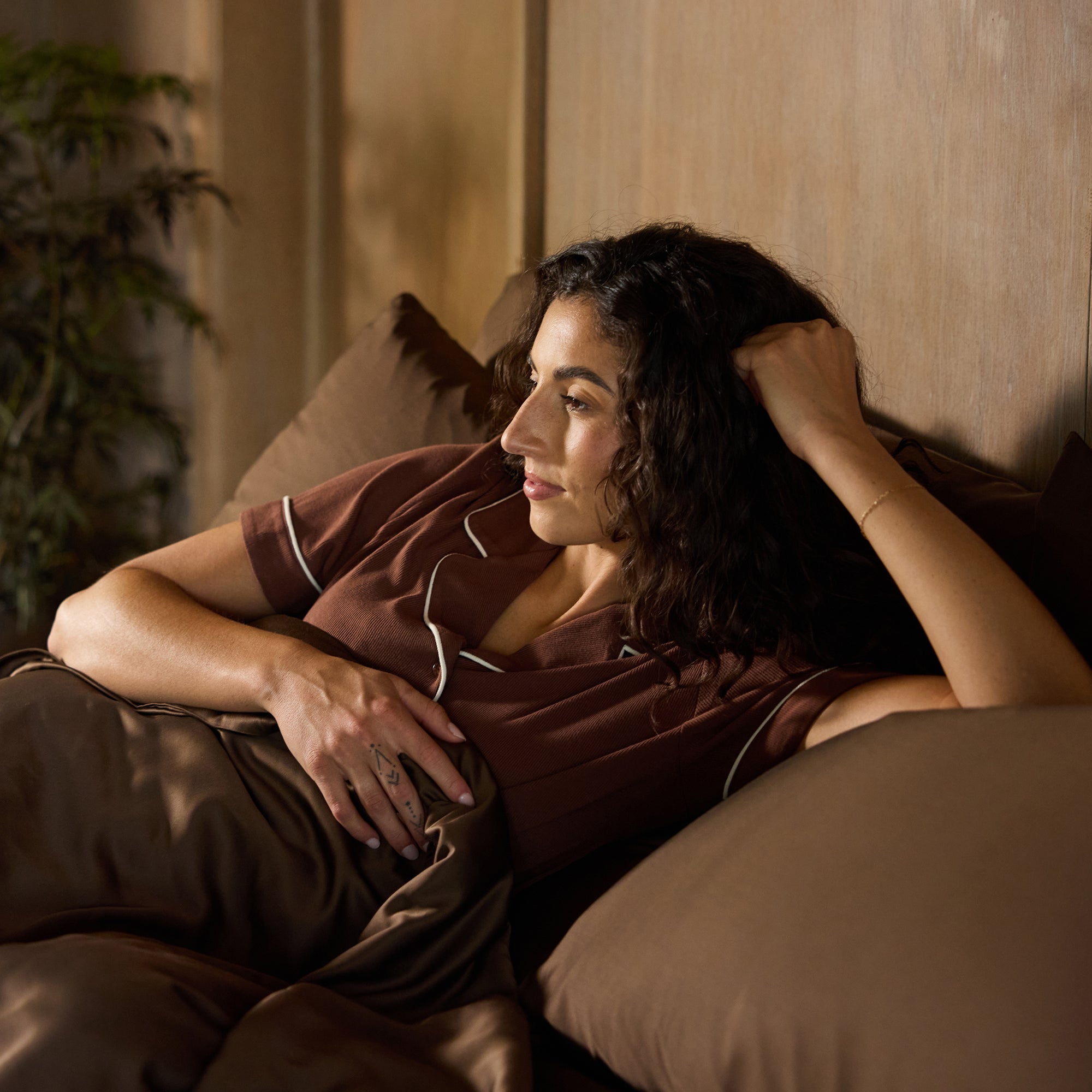 A woman with curly dark hair is lying in bed, resting her head on her arm. She's wearing brown pajamas and gazing off to the side thoughtfully. The bed is made with Cozy Earth's Bamboo Sheet Set, and there's a plant in the background. 